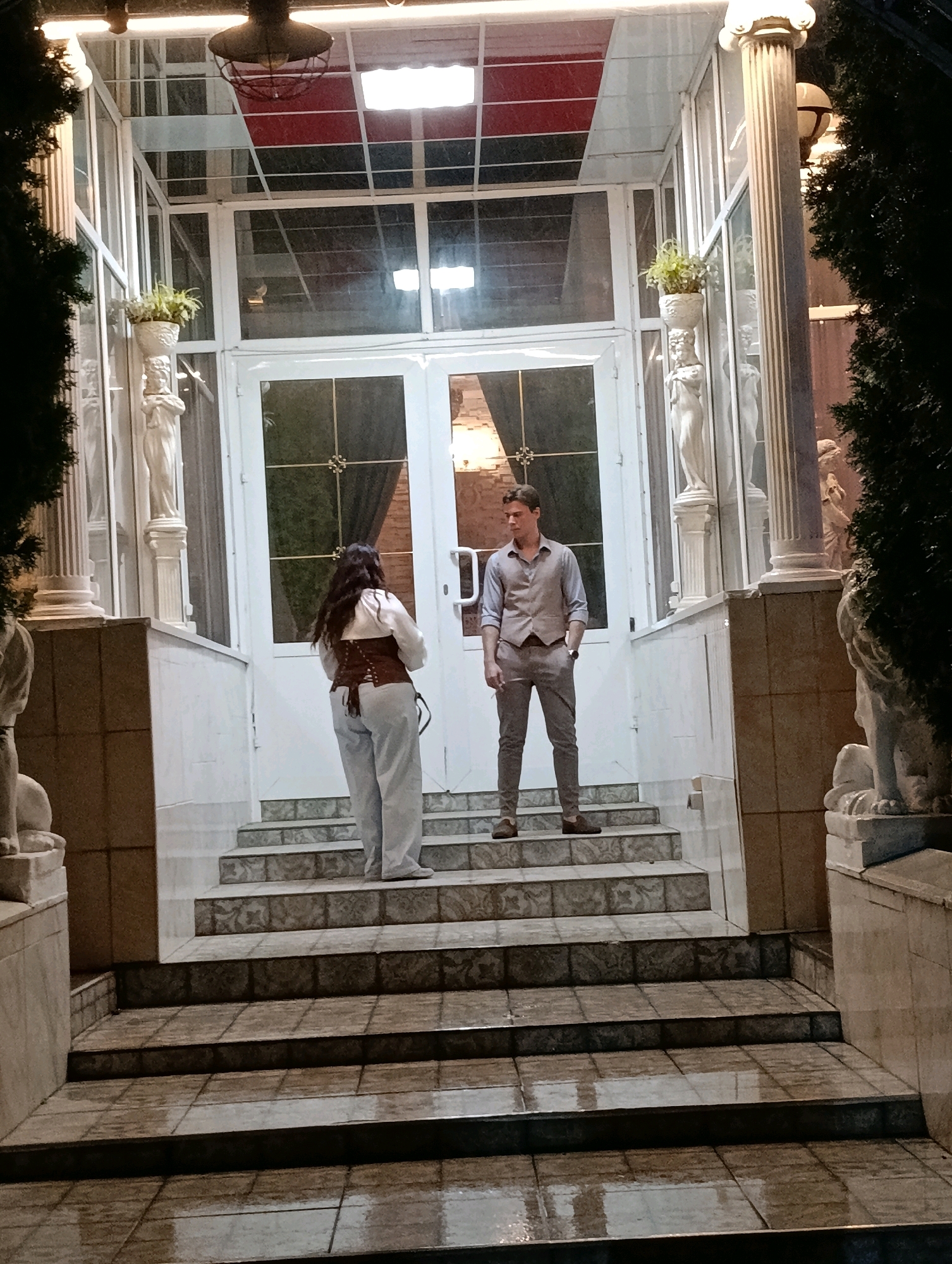 In the city of Korzhevo, Moldova, two individuals stand on a set of stone steps leading up to a glass-enclosed entranceway. The entrance has double doors and is flanked by decorative columns and statues holding potted plants. A man, dressed in a vest and dress pants, stands on the steps with his hands in his pockets, looking towards the woman. The woman, wearing a corset and loose pants, faces away from the camera, looking towards the doors. The steps and surrounding area appear to be wet, suggesting recent rain. The scene is illuminated by overhead lights and ambient exterior lighting.