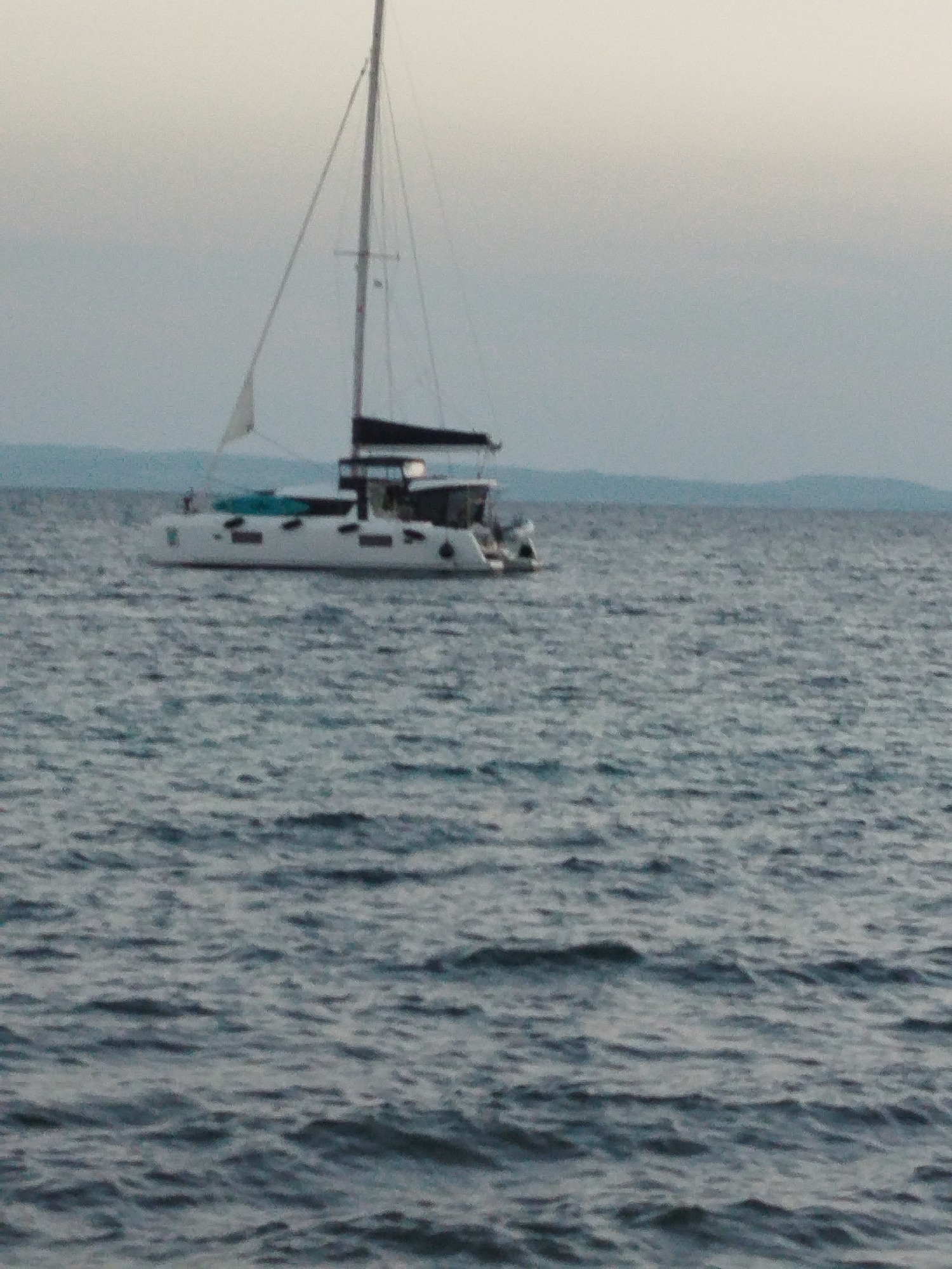 A white catamaran sailboat is on the water with a black sail. The sky is overcast and the water is choppy. In the distance, there is a faint outline of a coastline with hills. The scene appears to be in the sea near Sithonia, Greece.