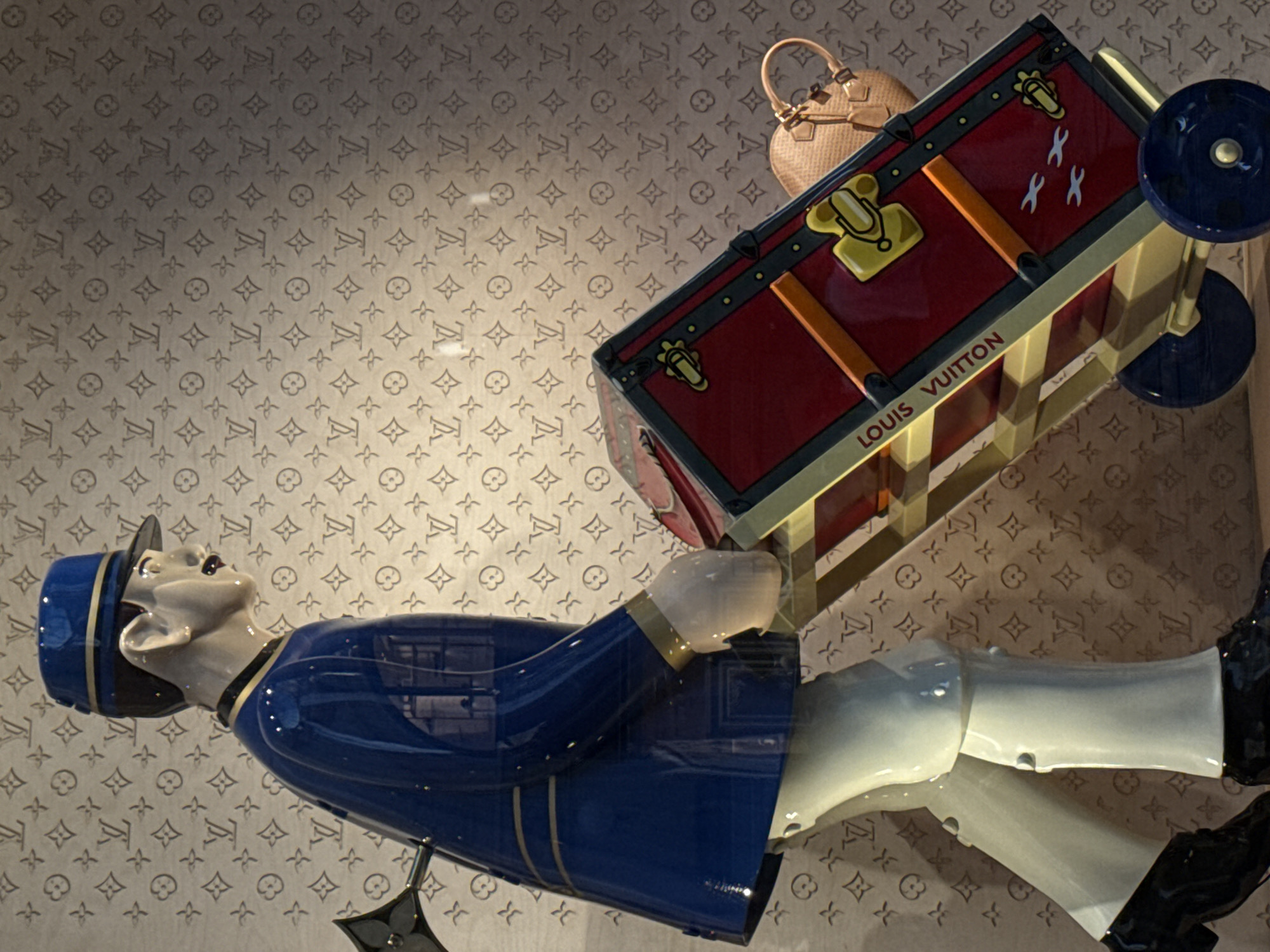 A display in Milan, Italy, shows a mannequin of a porter pulling a red and gold Louis Vuitton trunk, next to a tan purse, against a background patterned with Louis Vuitton logos. The porter, dressed in a blue uniform with white pants and black shoes, faces upwards. The trunk has black trim and gold hardware, with "LOUIS VUITTON" printed on its side. The background is a patterned wall or display panel.