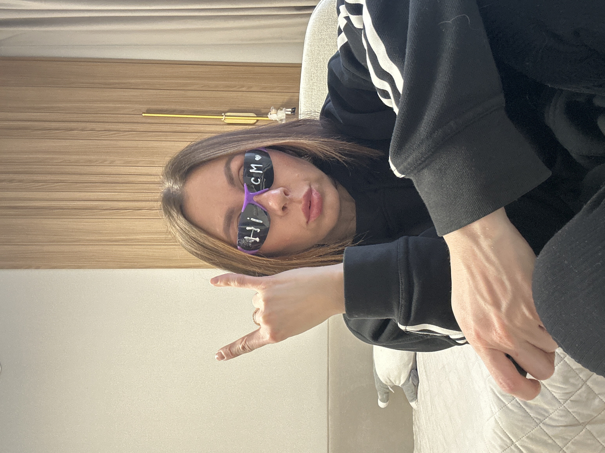A woman is posing for a photo indoors. She's wearing a black hooded sweatshirt with white stripes on the sleeves and sunglasses with the words "hi CM" written on the lenses. She is making the "rock on" hand gesture. The background includes a light-colored wall, a wooden-paneled area, and bedding. The location is not discernible as Votkinsk, Russia from the visual data provided.