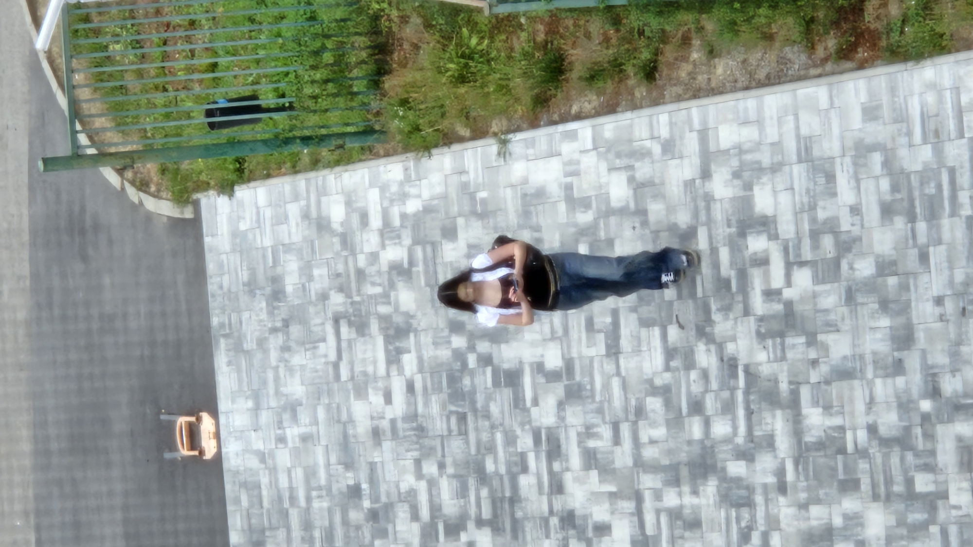 A young woman with long dark hair, wearing a white t-shirt over a dark tank top and blue jeans, walks on a paved path. She is looking at her phone. A dark backpack is slung over her right shoulder. In the background, a green metal fence borders a grassy area. To the left of the paved path, there is a grey asphalt surface with an orange object that appears to be a child's toy. The scene is an outdoor urban environment. The provided context suggests the location is Prešov, Slovakia.