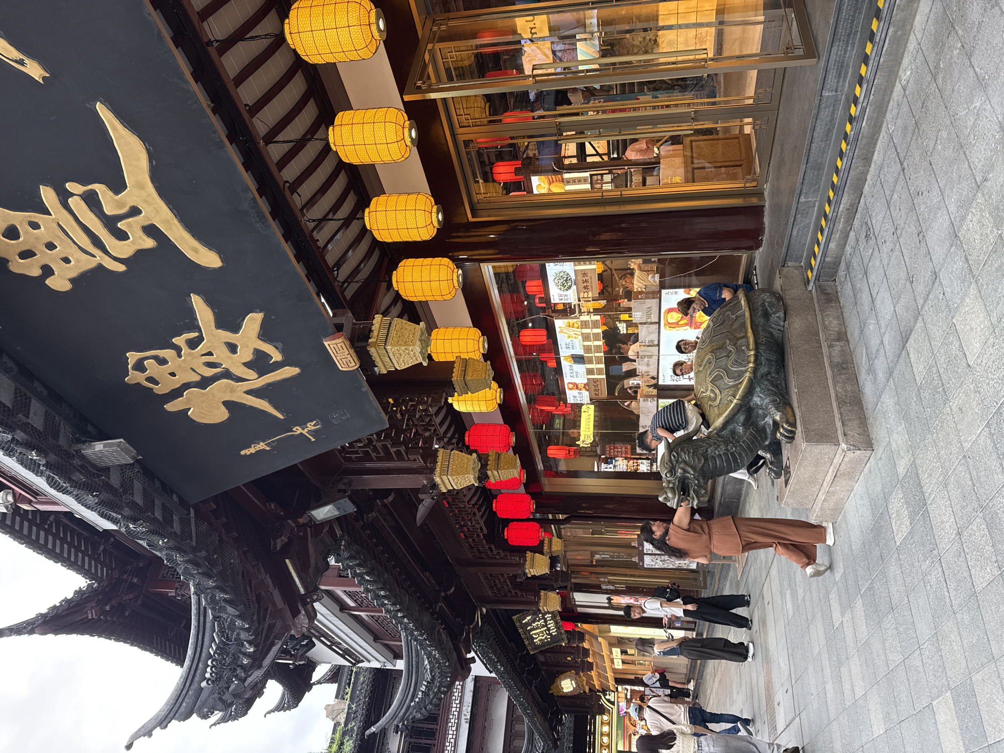 This image captures a bustling scene in front of a traditional Chinese building, likely a restaurant or shop, in Shanghai, China. The architecture features a distinctive upturned roof with ornate details and is adorned with numerous decorative lanterns, some red and some yellow. A large, golden Chinese character signage is prominently displayed above the entrance.
In the foreground, a large bronze sculpture of a turtle stands on a stone pedestal. A woman in a brown outfit is seen interacting with the sculpture, while a child in a striped shirt is perched on its back. Several other people are walking on the paved walkway in the background, some appearing to be tourists or shoppers. The storefront has large glass doors and windows, revealing glimpses of the interior, which appears to be a commercial establishment with displays and seating. The overall atmosphere suggests a lively street scene in a well-trafficked area of Shanghai, possibly a historical district or a popular tourist destination.
