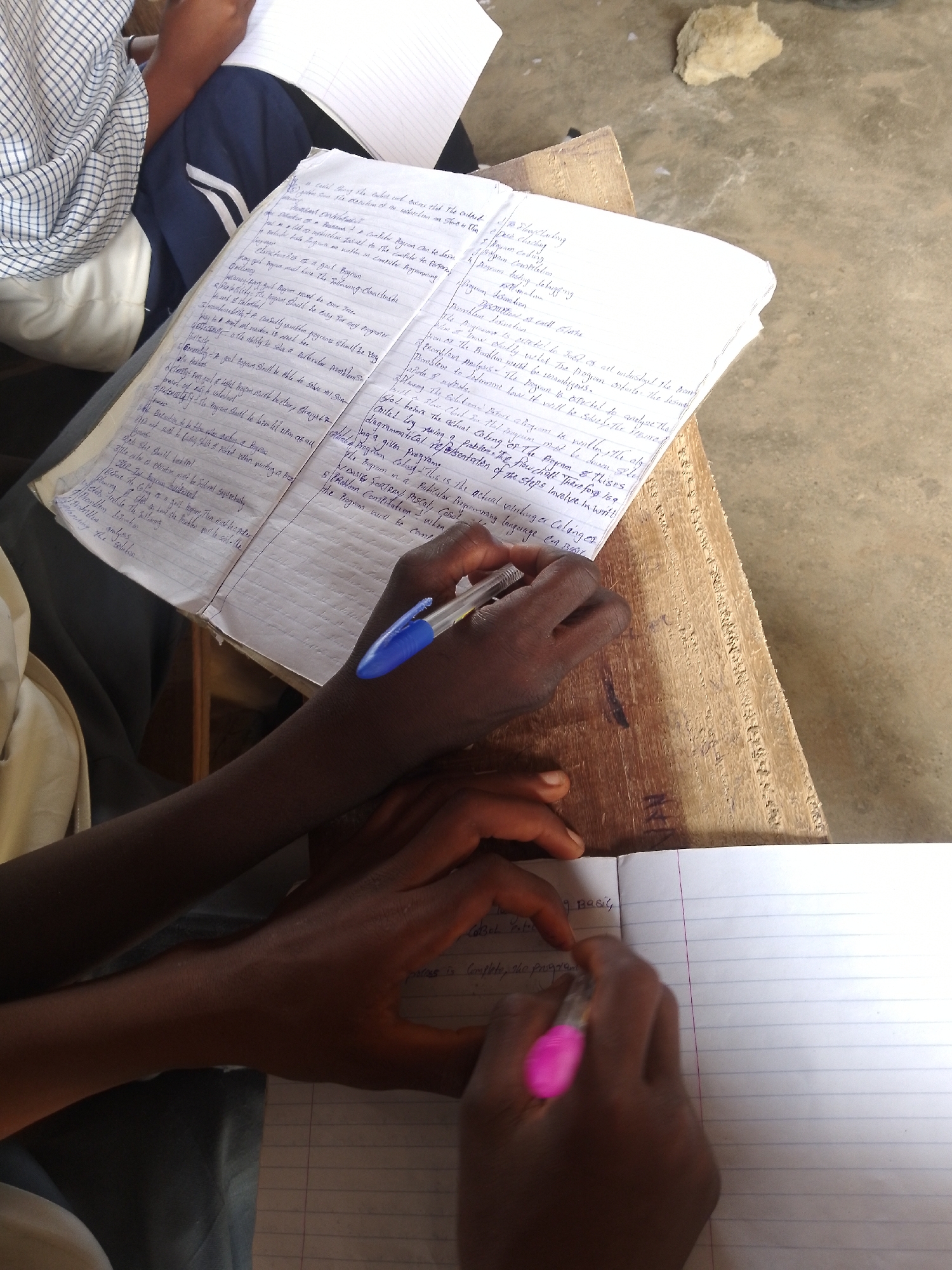 This media file captures a close-up view of two individuals engaged in writing within notebooks, suggesting an academic or study setting. The foreground is dominated by the hands and arms of two people, one with a blue pen and the other with a pink pen, actively writing in lined notebooks. The notebooks are open, revealing handwritten text, possibly notes or assignments. The larger notebook, in the upper half of the image, is filled with dense handwritten text. The smaller notebook, in the lower half, also has handwritten text, but more of its pages are visible and appear to be in progress. The scene is lit by natural light, possibly indicating daytime. The background is somewhat out of focus, showing a wooden surface that appears to be a table or desk, and a hint of a textured surface possibly a wall or floor. A light-colored, rough object, perhaps a stone or piece of plaster, rests on the wooden surface in the upper right. The overall atmosphere is one of focused activity and learning. The visible text in the notebooks appears to be in English and contains technical terms related to programming, such as "program," "coding," "debugging," "compilation," and "flowchart." There are also numbers and bullet points, indicating structured notes. The individuals' hands suggest varying skin tones.