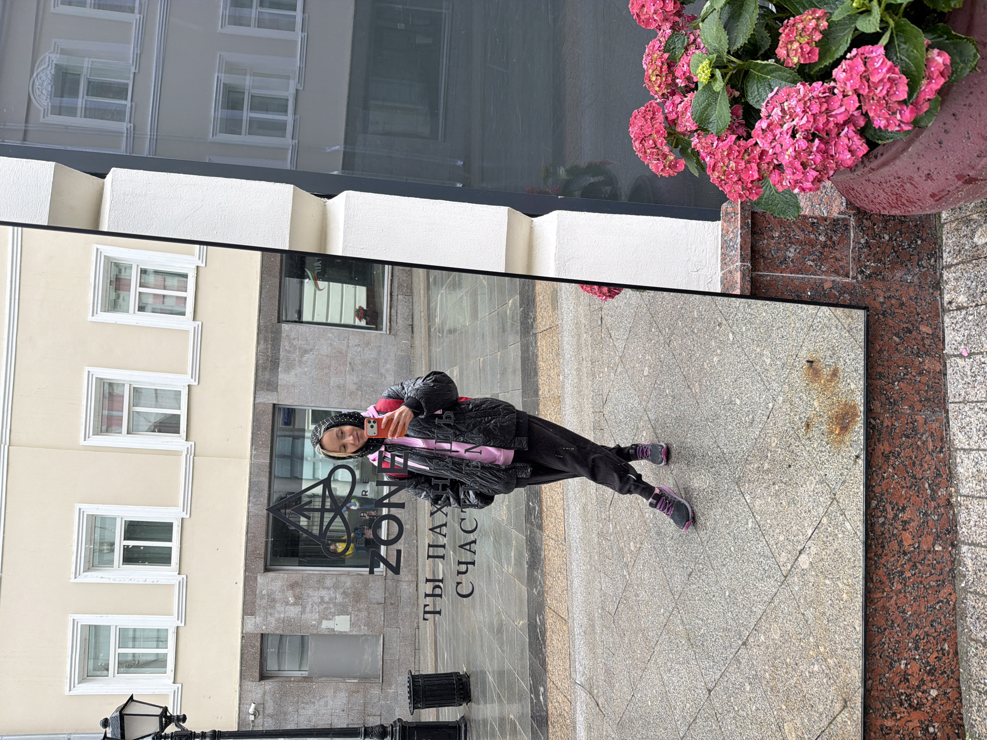 The image depicts a woman taking a selfie in a reflective surface in Moscow, Russia. The background reflects buildings with a combination of light yellow and gray facades and windows. In the foreground, there is a flower pot with pink hydrangeas on a red granite base. The woman is wearing a black jacket, black pants, and gray/purple sneakers. She is holding a phone in her hand. The reflection includes a street with decorative lamp posts and the lettering "ZONE Ты ПАХАЕТ СЧАСТЬЕ" which is the name of the restaurant Zone and translates to "You farm happiness". The reflection shows the cityscape and a person taking a photograph in the mirror.
