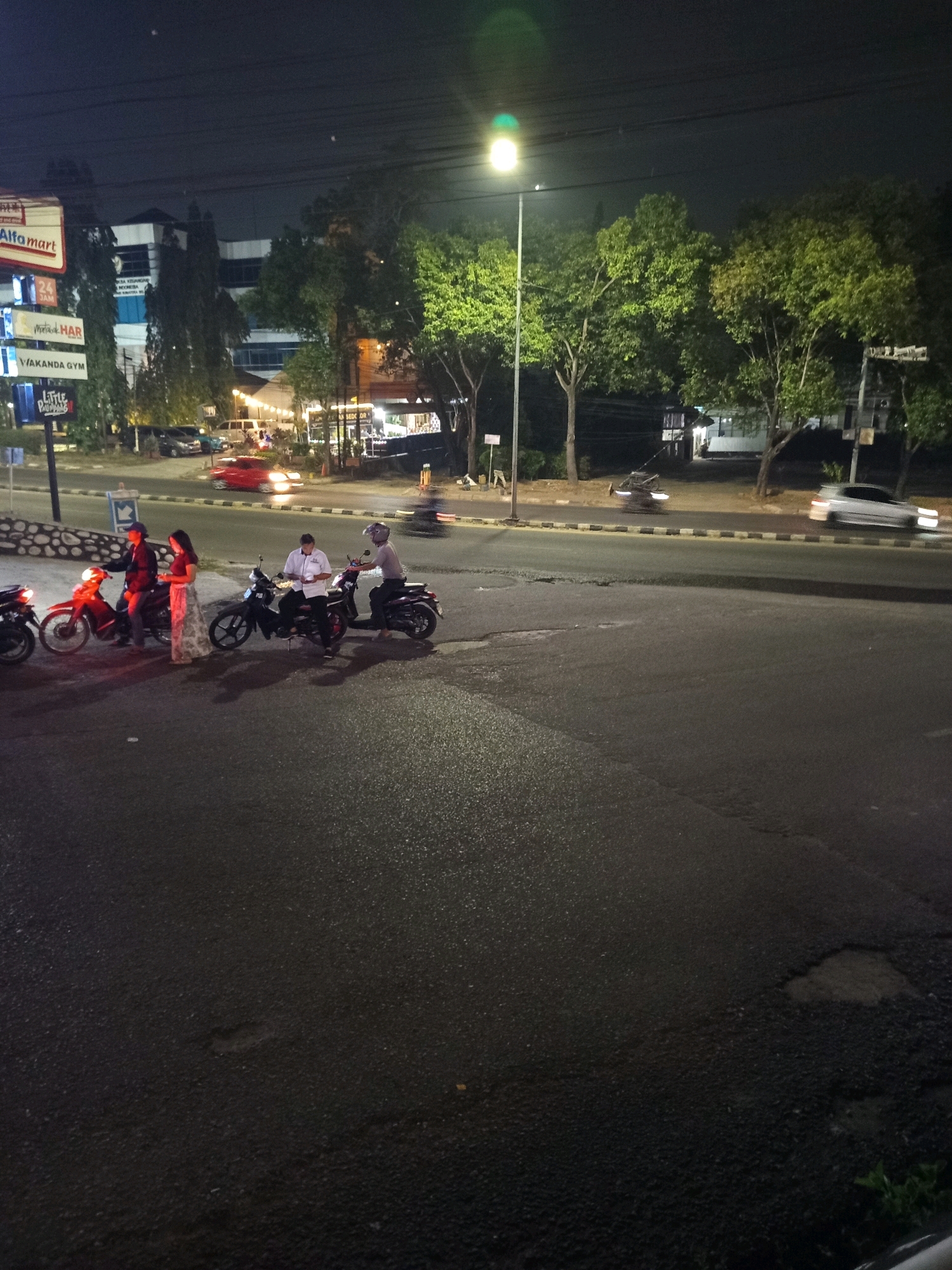 At night on a street in Palembang, Indonesia, several people are gathered around motorcycles. The scene is illuminated by a street lamp and the lights from nearby buildings, including an Alfamart.
In the foreground, three motorcycles are parked on the side of the road. Two people are standing next to a red motorcycle, and two other individuals are seated on black motorcycles. One of the individuals on a black motorcycle appears to be wearing a white shirt and holding something.
In the background, cars are driving on the road, with one red car blurred by motion. A white car is also visible further down the street. Trees line the side of the road, and various signs are displayed on buildings. The overall environment suggests a commercial or semi-urban area.