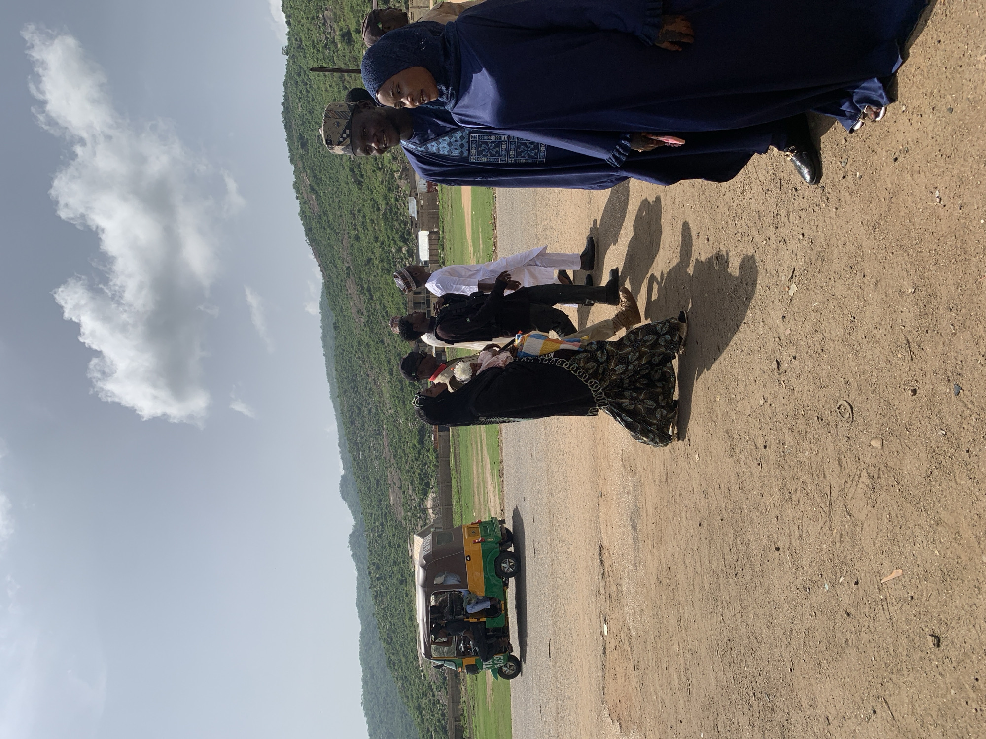 The image depicts a group of people standing on a dirt road in what appears to be Naraguta Hausawa, Nigeria. The terrain is arid, and there is a mountain in the background. A tuk-tuk sits parked on the road. The people are wearing a variety of clothing, including traditional attire.