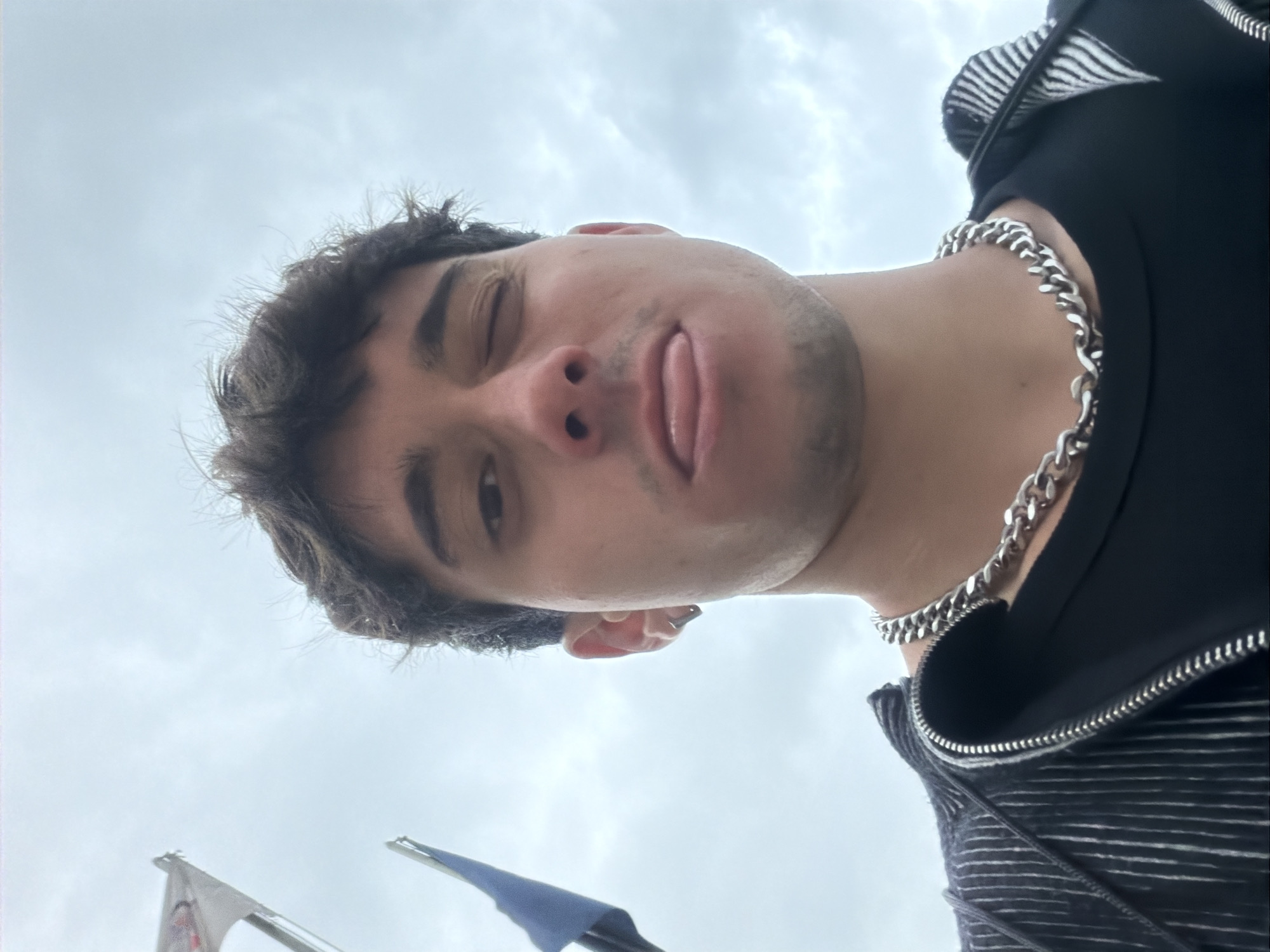 A low-angle, close-up photograph captures a young male individual from a worm's-eye perspective against an overcast sky. The subject, with dark, curly hair, is looking directly towards the camera, winking his left eye (viewer's right) and playfully sticking his tongue out. He is wearing a dark top, possibly a jacket, featuring a zipper and a striped pattern on a visible inner layer or collar. A prominent, thick silver-colored chain necklace adorns his neck. The background consists solely of a bright, cloudy sky, suggesting an outdoor setting during the daytime with diffused light. Towards the bottom left corner of the frame, parts of two flagpoles with flags are visible; one flag is white with indistinct details, and the other appears to be blue. The overall mood conveyed by the subject's expression is playful and casual. There is no visible text in the image. Given the context, the setting is Sofia, Bulgaria.