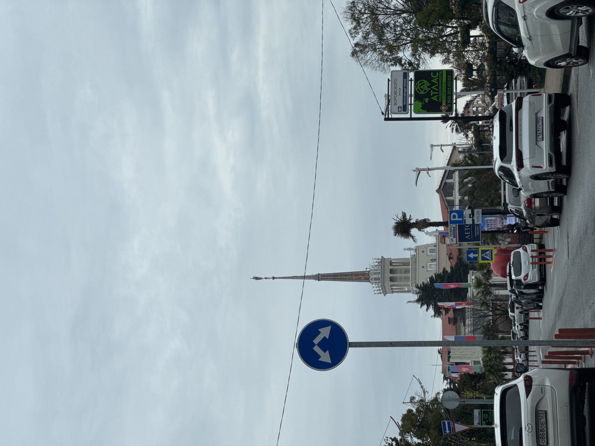 The image captures a street scene in Sochi, Russia, characterized by a mix of urban elements and architectural landmarks. A blue circular sign with dual directional arrows is prominently featured in the lower center, suggesting a fork in the road. The spire of the Sochi Marine Station stands in the background. A street sign reads "Vorovskogo street" in both Russian and English. A green advertisement for real estate company ATLAC and the street name POYARKS appear. Several white cars are parked along the street, with license plates visible. Flags are mounted to flagpoles. The sky is overcast.