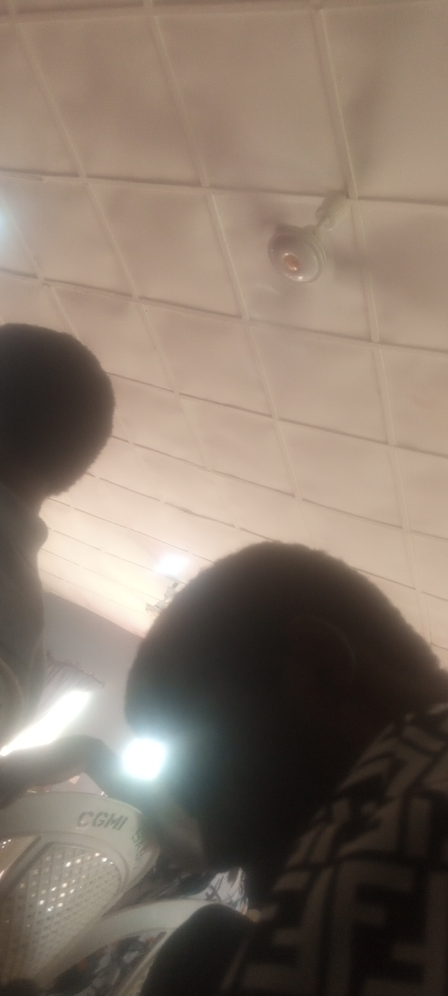 The image, likely taken indoors in Sabon Tasha, Nigeria, shows a low-angle view of a tiled ceiling with a ceiling fan. Two people are partially visible in the foreground: one on the left with an afro-like hairstyle and another on the right with a similarly styled hair. A bright light source, possibly a phone's flashlight, is held near the left person. A white plastic chair with the letters "CGMI" is also visible. The focus is poor, and the lighting is uneven.