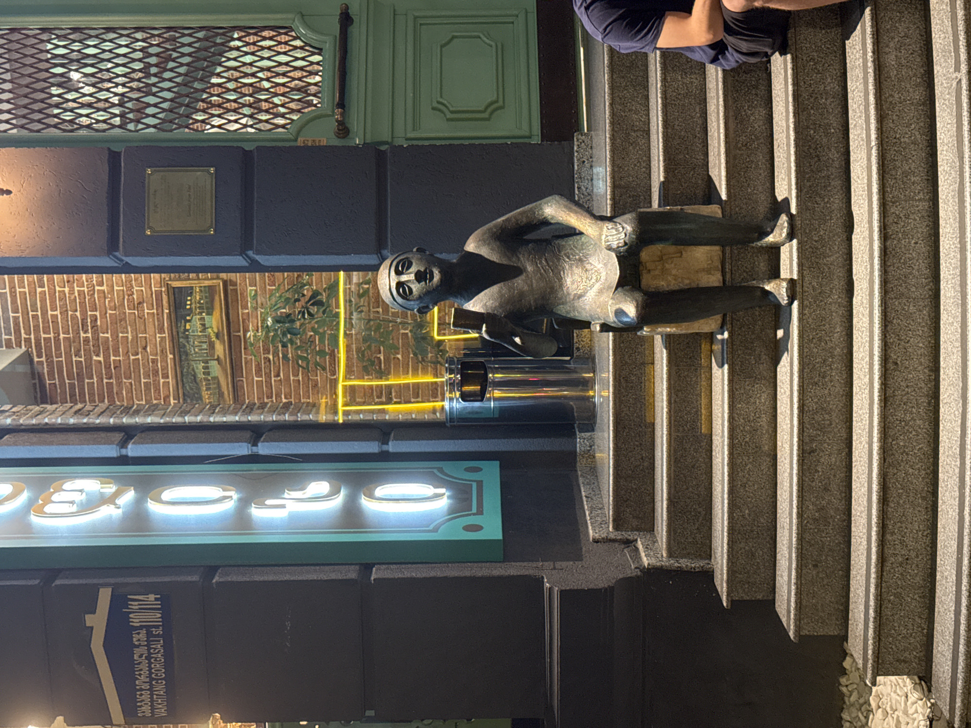 The image depicts a street scene in Batumi, Georgia, focusing on the entrance of a building with an illuminated sign and a bronze statue.
In the foreground, a bronze statue of a seated person holding a small object is positioned on the steps leading up to the building. The statue is weathered and appears to be a public art installation. To the right of the statue, the legs of a person wearing a dark blue t-shirt and dark shorts are visible, also seated on the steps.
The building entrance features a dark grey facade with a bright green double door on the right, adorned with decorative metalwork. Above the door, there is a plaque and a framed painting on the brick wall. To the left of the door, a vertical sign with illuminated letters, appearing to spell out "B", "e", "c", "o", "m", "e", is mounted on the dark facade. Below the sign, a street address plate reads "VAKHTANG GORGASALI st. 110/114" in English and Georgian. A modern stainless steel trash bin with a black lid is placed to the left of the statue.
The steps leading up to the entrance are made of granite with contrasting dark strips on the edges. The ground to the left of the steps is covered with white pebbles. The overall lighting suggests it is evening or nighttime, with artificial lights illuminating the scene. The environment appears to be urban, likely a commercial or residential area.