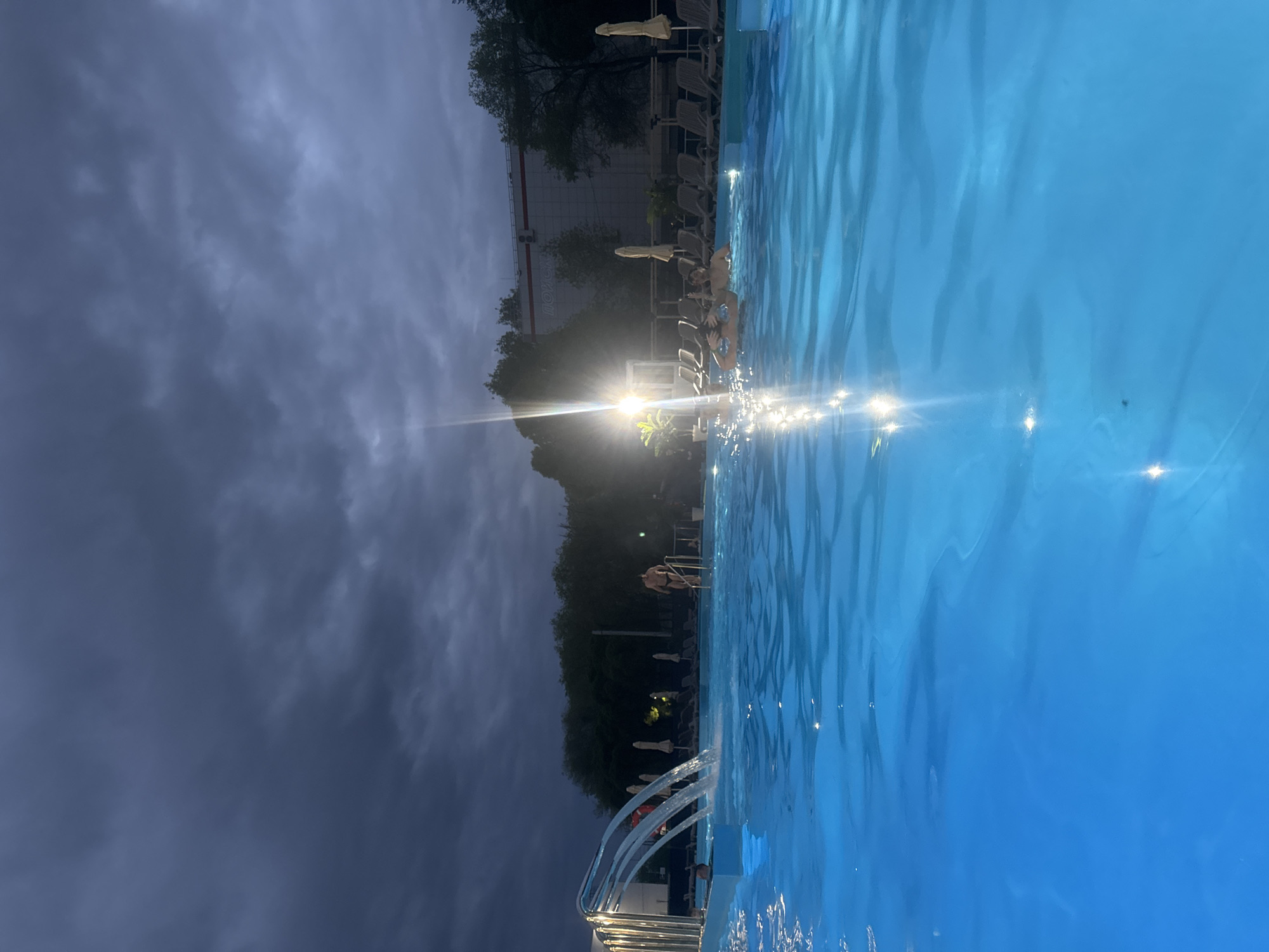 The image shows a swimming pool located outdoors with dark, stormy clouds overhead. The sky is divided into two distinct parts: the left side is dark grey with heavy clouds, while the right side is lighter, though still overcast. A bright light source is visible in the distance, partially obscured by trees. Along the side of the pool, there are numerous lounge chairs with white umbrellas, some appearing to be folded down. People are visible near the pool, some seemingly in the water. A metal ladder leads into the pool. Water from the pool appears to be overflowing in a cascade or fountain-like feature near the ladder. The photograph is likely taken at or near the hotel Imperial Club Deluxe in Ulyanovsk, Russia.
