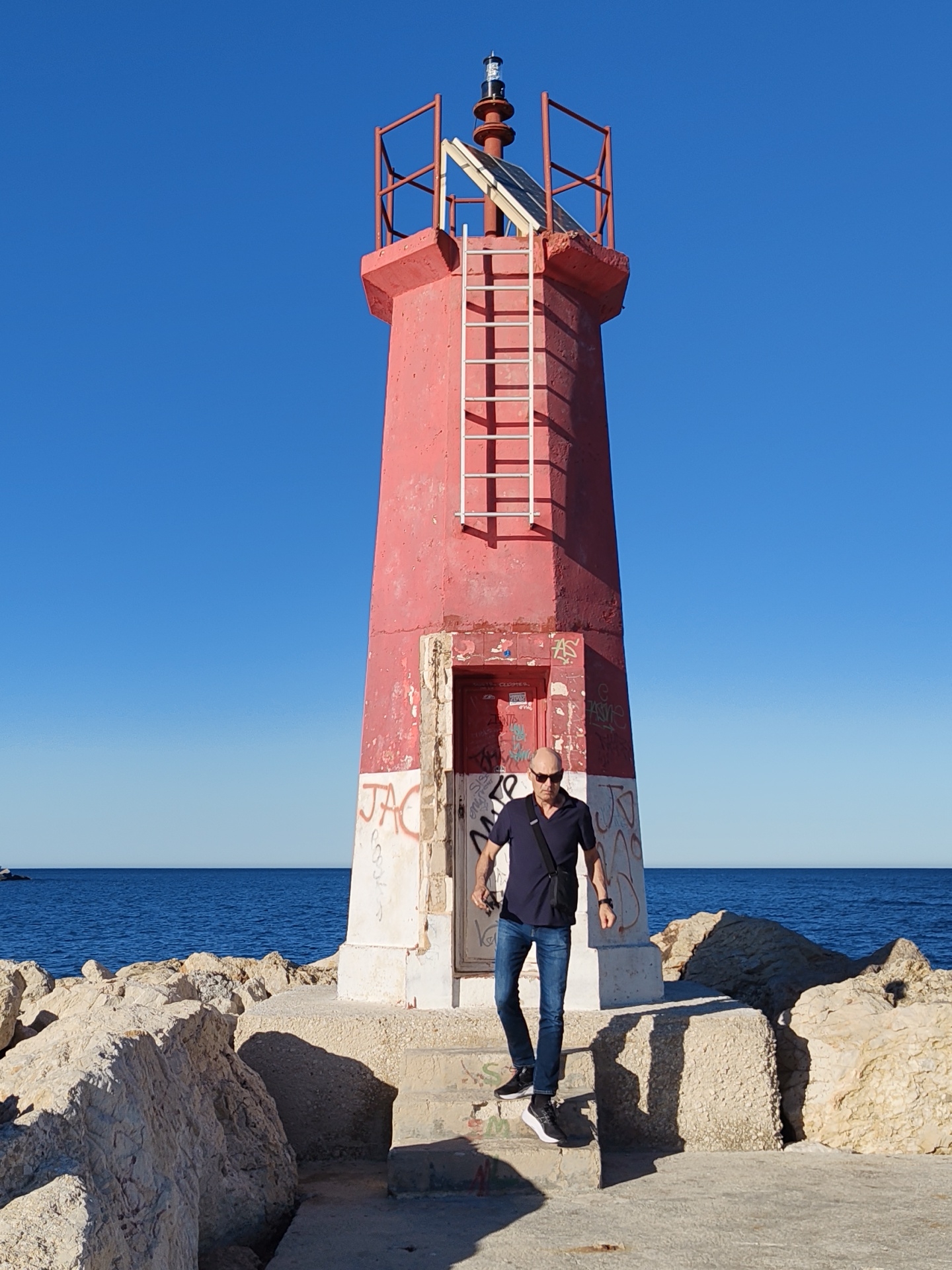 A full-length outdoor shot captures a vibrant red and white lighthouse on a sunny day in Dénia, Spain. The small beacon tower is situated on a concrete and rocky breakwater, with the deep blue sea stretching behind it under a clear, cloudless sky. The lighthouse's base is painted white and is heavily adorned with red and black graffiti, prominently featuring "JAC" in red on the left side. A white ladder ascends one side of the red structure to a platform equipped with a solar panel and the light fixture at the very top.
In the foreground, a man is walking down concrete steps away from the lighthouse, towards the left of the frame. He is bald, wears dark sunglasses, a dark blue polo shirt, blue jeans, and black sneakers, with a black shoulder bag slung across his body. The bright sun casts distinct, elongated shadows of the man and the lighthouse on the concrete surface, suggesting it is either morning or late afternoon. The immediate surroundings include large, light-colored rocks forming the breakwater, contrasting with the vibrant sea. Some green graffiti is also visible on the concrete steps near the man's feet. The scene depicts a calm, clear day at a coastal location.