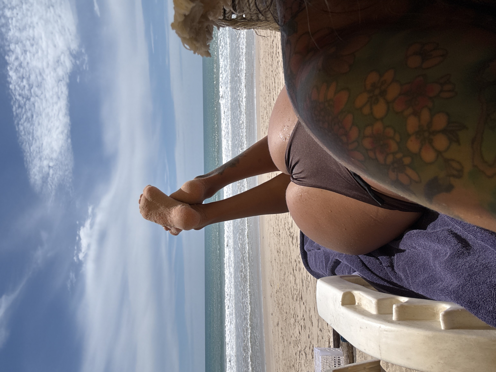 A first-person perspective shot shows a person lying on a lounge chair on a sandy beach with the ocean and sky in the background. The person is wearing brown bikini bottoms. Their feet, covered in sand, are crossed and raised, with the soles visible. A portion of the person's tattooed arm is in the foreground, displaying a floral pattern. A purple towel is draped over the lounge chair. To the left of the frame, a white plastic basket is visible. The ocean has white waves breaking on the shore, and the sky is blue with scattered white clouds. The scene appears to be in Choeng Thale, Thailand.