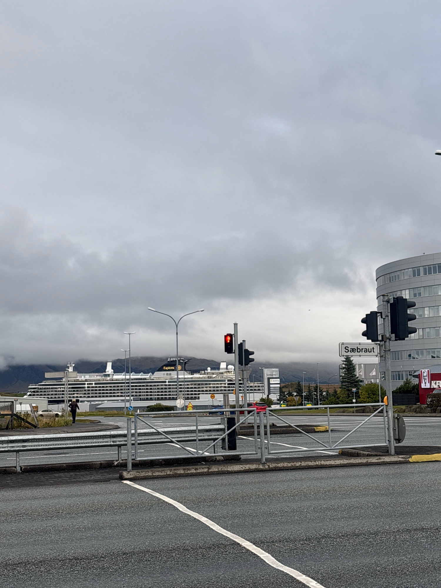The image captures a view of Reykjavik, Iceland, with a large cruise ship docked in the background. The sky is overcast and cloudy. In the foreground, there is a street intersection with traffic lights, one of which is red. A road sign reading "Sæbraut" is visible. To the right, a modern, multi-story building with a rounded facade stands. A KFC restaurant with its distinctive red and white signage is also present on the right side of the image. In the mid-ground, a person is walking away from the camera towards the cruise ship. The cruise ship itself is white with multiple decks and appears to be named "Explorers." Mountains shrouded in clouds are visible behind the ship. The scene depicts a port area with urban elements and natural landscape features.