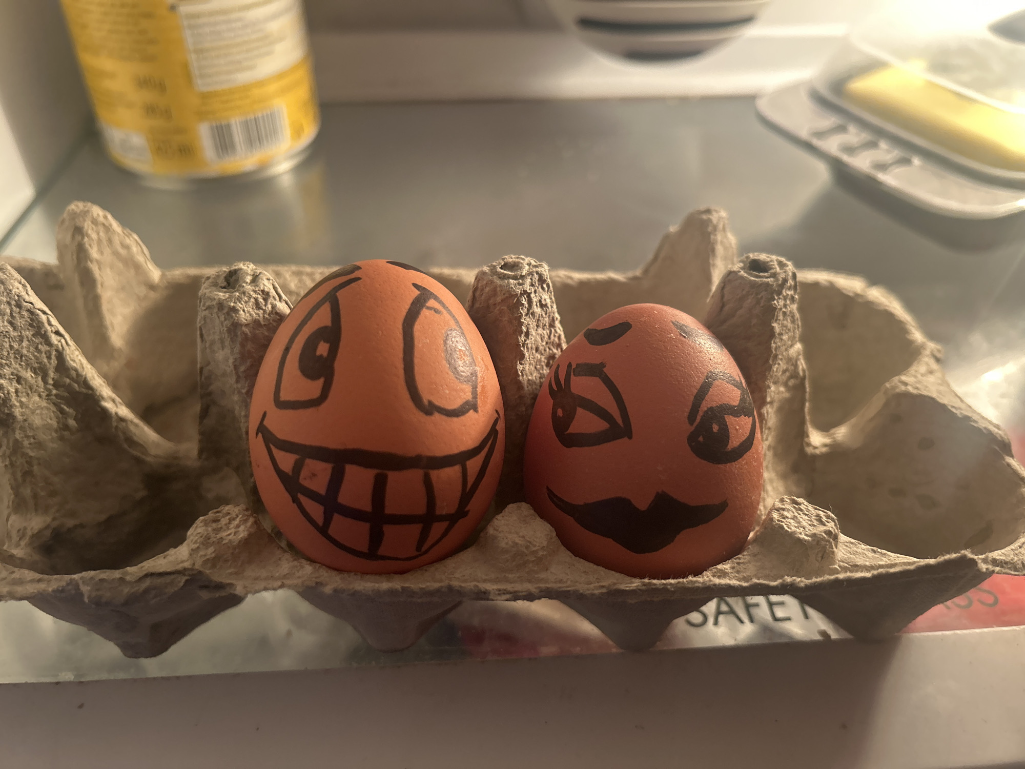 The image shows a carton of eggs inside a refrigerator. Two of the eggs have faces drawn on them in black marker. The egg on the left has a wide, toothy grin and large, round eyes. The egg on the right has one eye winking and a large, curved smile. The eggs are in a cardboard egg carton labeled "SAFE". Behind the eggs, on the refrigerator shelf, there is a yellow container, a dish with some food in it, and a butter dish with butter in it. The location is unknown.