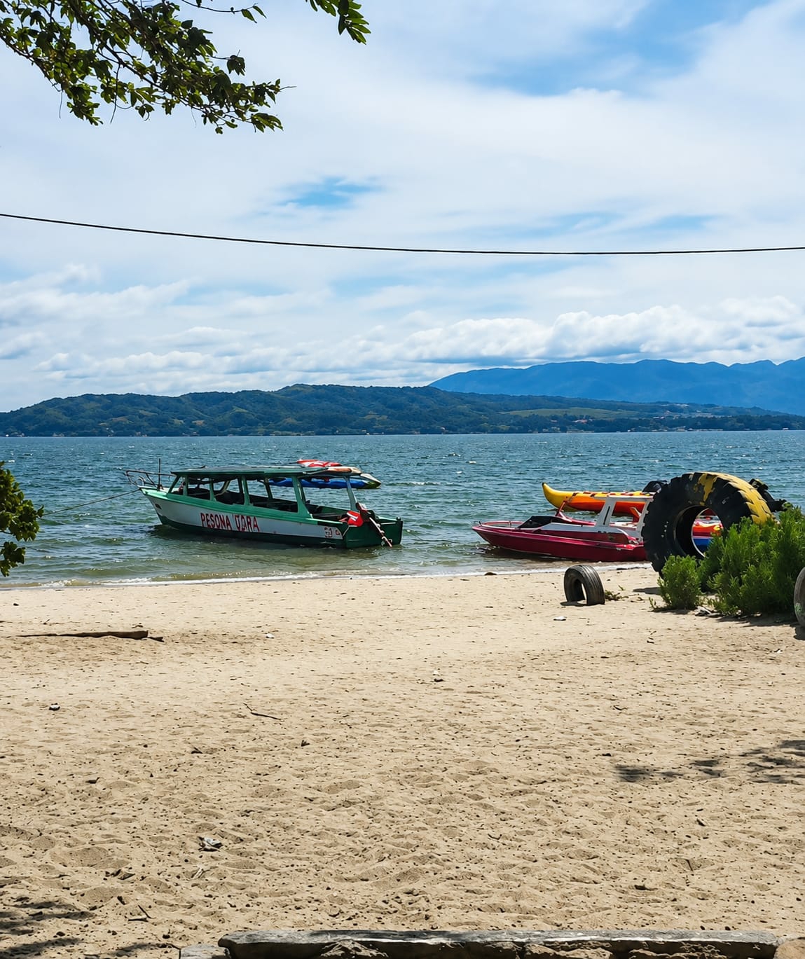 The image depicts a serene lakefront scene in Lumban Bul Bul, Indonesia. In the foreground, a sandy beach stretches towards the water, with scattered driftwood. The calm lake is dotted with several boats, including a green and white passenger boat labeled "PESONA TUARA" and a smaller red boat with a yellow kayak on top. In the background, lush green hills rise against a backdrop of a cloudy blue sky. A tree branch with green leaves hangs from the top left corner of the frame, adding a touch of nature to the composition. Several large tires are visible on the shore, suggesting a recreational area.