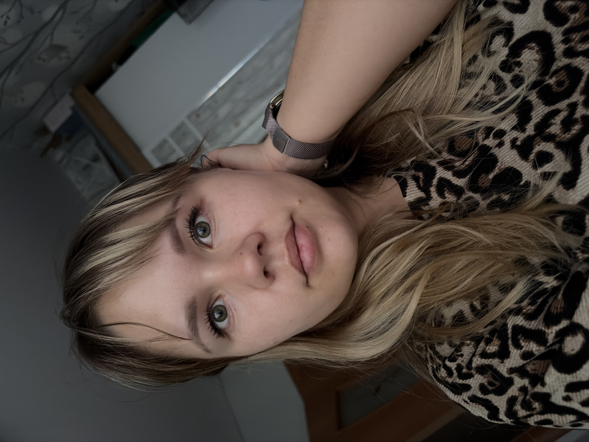 A close-up, tilted portrait of a young woman with fair skin and blonde hair. She is wearing a leopard-print shirt. Her left hand is raised to her head, and a gray watch is visible on her wrist. The background is blurred, with a glimpse of a white shelf and patterned wallpaper. The location is not discernible from the image.