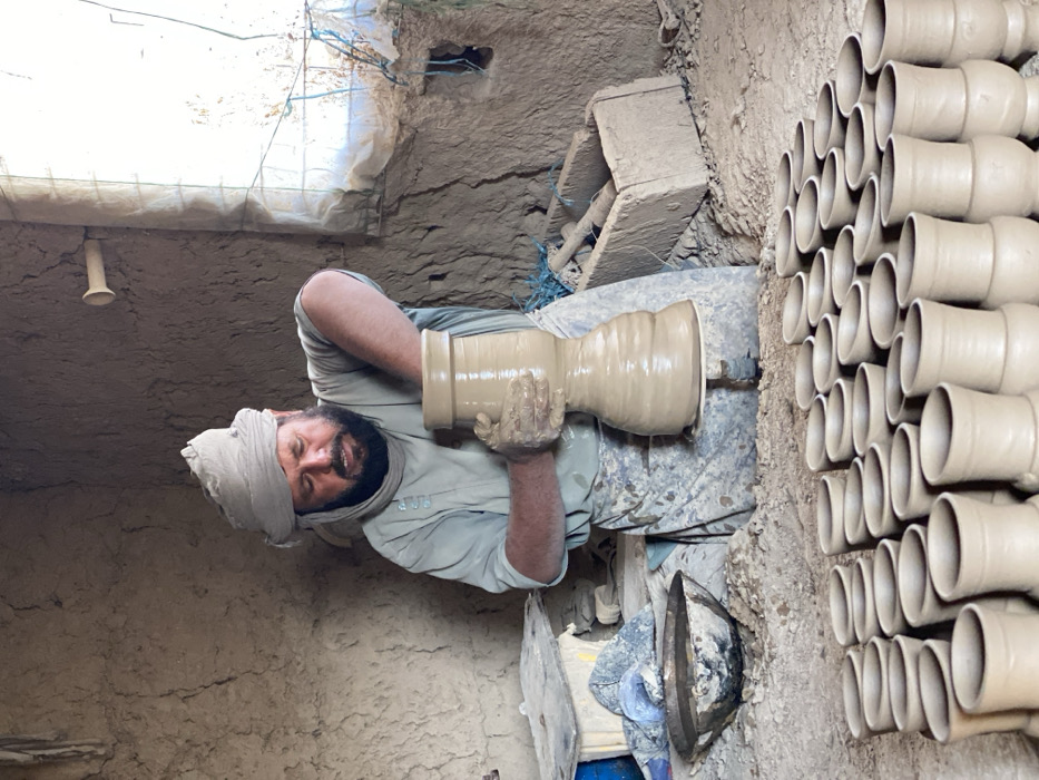 An adult male artisan is depicted within a rustic, mud-walled pottery workshop in Kandahar, Afghanistan. He wears a light-colored turban, a light-blue tunic, and a stained apron, and has a dark beard. The artisan is seated at a potter's wheel, actively shaping a large, unglazed clay vessel with an hourglass-like profile, its surface visibly wet with clay. His hands are coated in clay. To his immediate left, a metal bowl filled with liquid or wet clay rests on a debris-strewn platform. In the foreground, numerous identically shaped, smaller, unglazed clay vessels are arranged in organized rows on the dirt floor, indicating a drying or storage area. The workshop walls are constructed from unplastered adobe or mud brick, showing cracks and irregular textures. A bright, rectangular window opening on the right is covered with a translucent plastic sheet secured by wires or string, serving as the primary light source. A wooden crate containing tools is visible on the floor behind the artisan, and the entire floor is covered in dried clay dust and debris. A small, light-colored cylindrical object protrudes from the upper right wall.