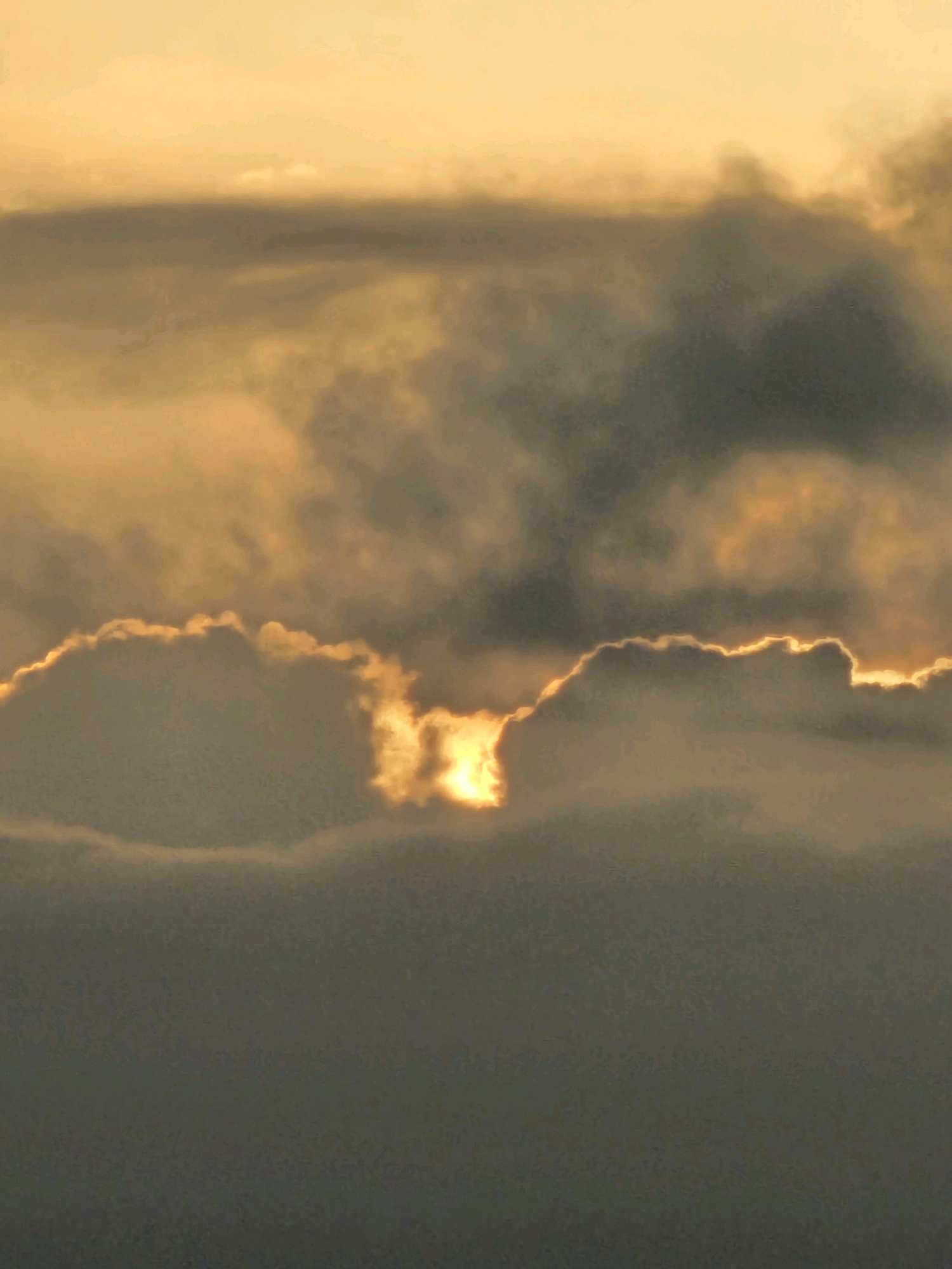 The image captures a dramatic sunset sky over Denpasar, Indonesia, with the sun partially obscured by thick, dark clouds. The lower portion of the sky is a deep, somber gray, indicative of heavy cloud cover. In contrast, the upper sky is illuminated by the setting sun, casting a warm, golden-yellow glow. This light brilliantly outlines the edges of the clouds, creating a striking contrast between light and shadow. The clouds themselves are varied in texture and density, with some appearing wispy and others dense and turbulent. There are no visible people, structures, or specific landmarks that would pinpoint a precise location within Denpasar, making it a generic depiction of a sunset over the city. The overall mood is one of awe and intensity, with the vibrant colors of the sunset battling against the encroaching darkness of the clouds.
