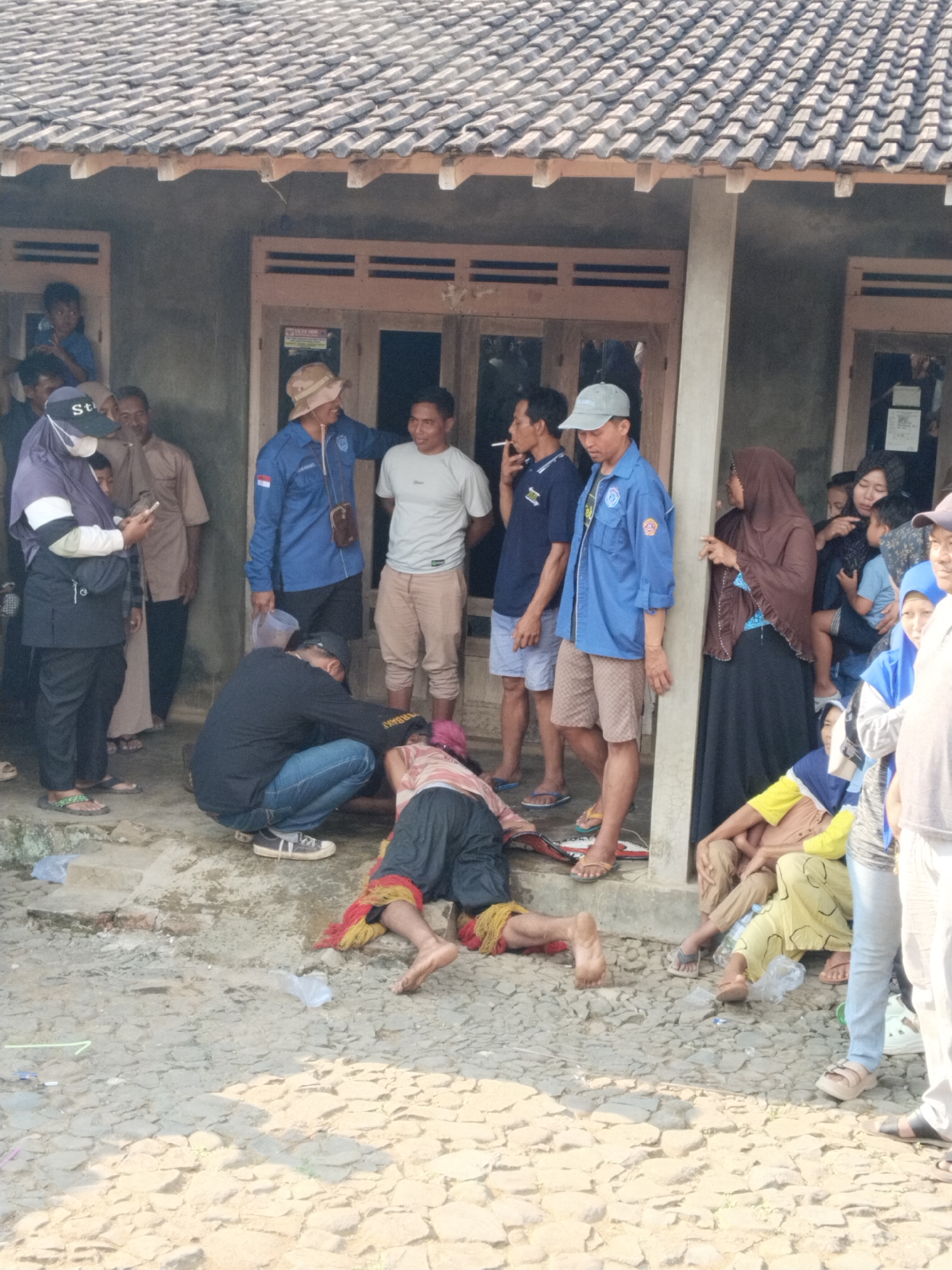 A group of people are gathered outside a building with a tiled roof in what appears to be a village setting in Ngadirejo, Indonesia. A man in a black shirt and blue jeans is kneeling down and tending to another person lying on the ground, who is wearing a red and yellow garment around their waist and dark pants. Several other individuals are standing and observing the scene, some are smoking. The ground is covered with rough, uneven stones and some plastic debris is scattered around. The building appears to be a simple house with wooden doors and windows. The scene suggests a public gathering or a situation where someone may have fallen or is receiving assistance.