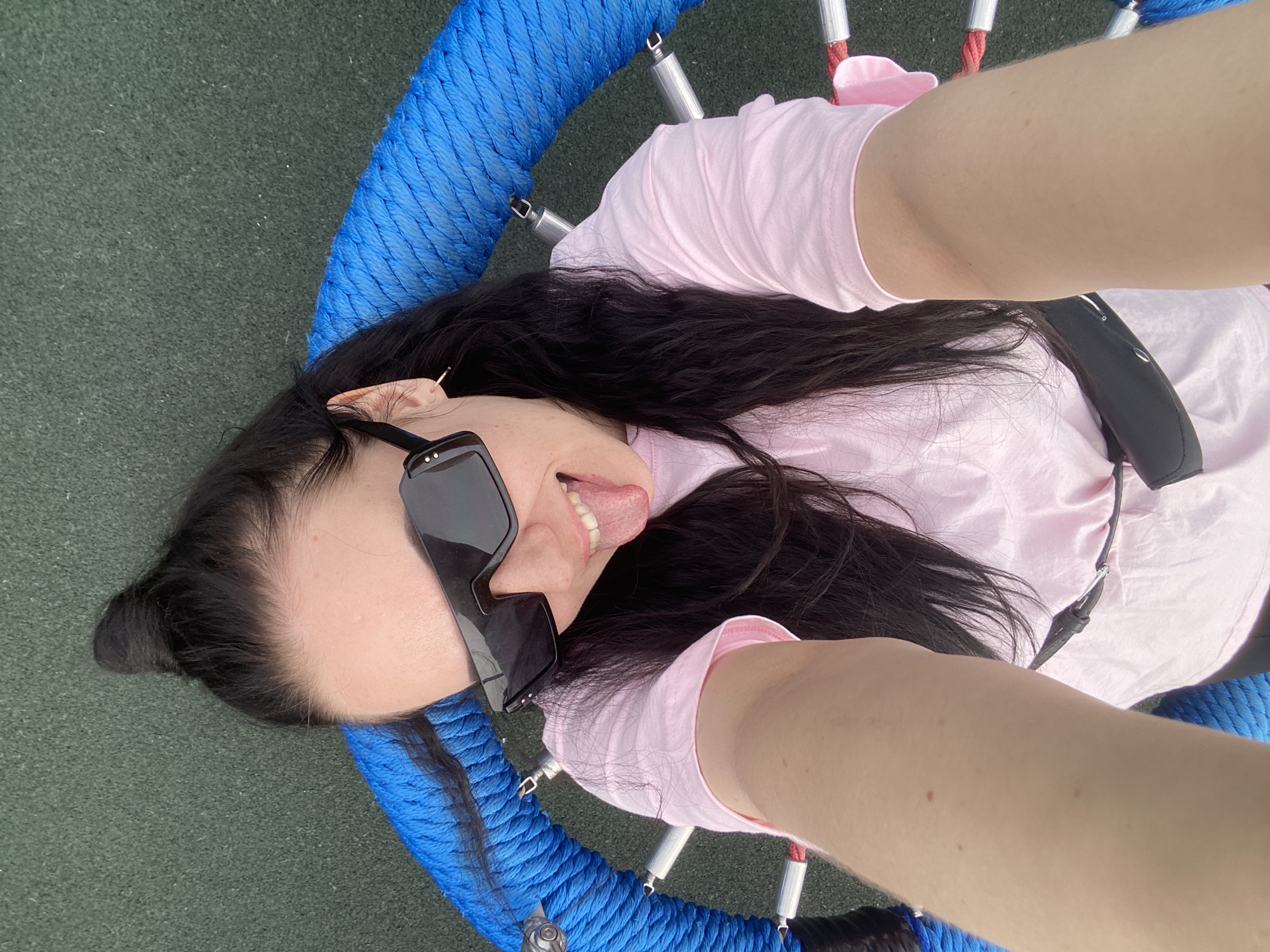 A woman lies in a blue, circular swing set at an outdoor playground. The ground is green. She wears a pink shirt and black sunglasses, and her tongue is sticking out. Her dark hair is pulled back into a bun. A black bag is also visible in the photo. The location is in Perm, Russia.