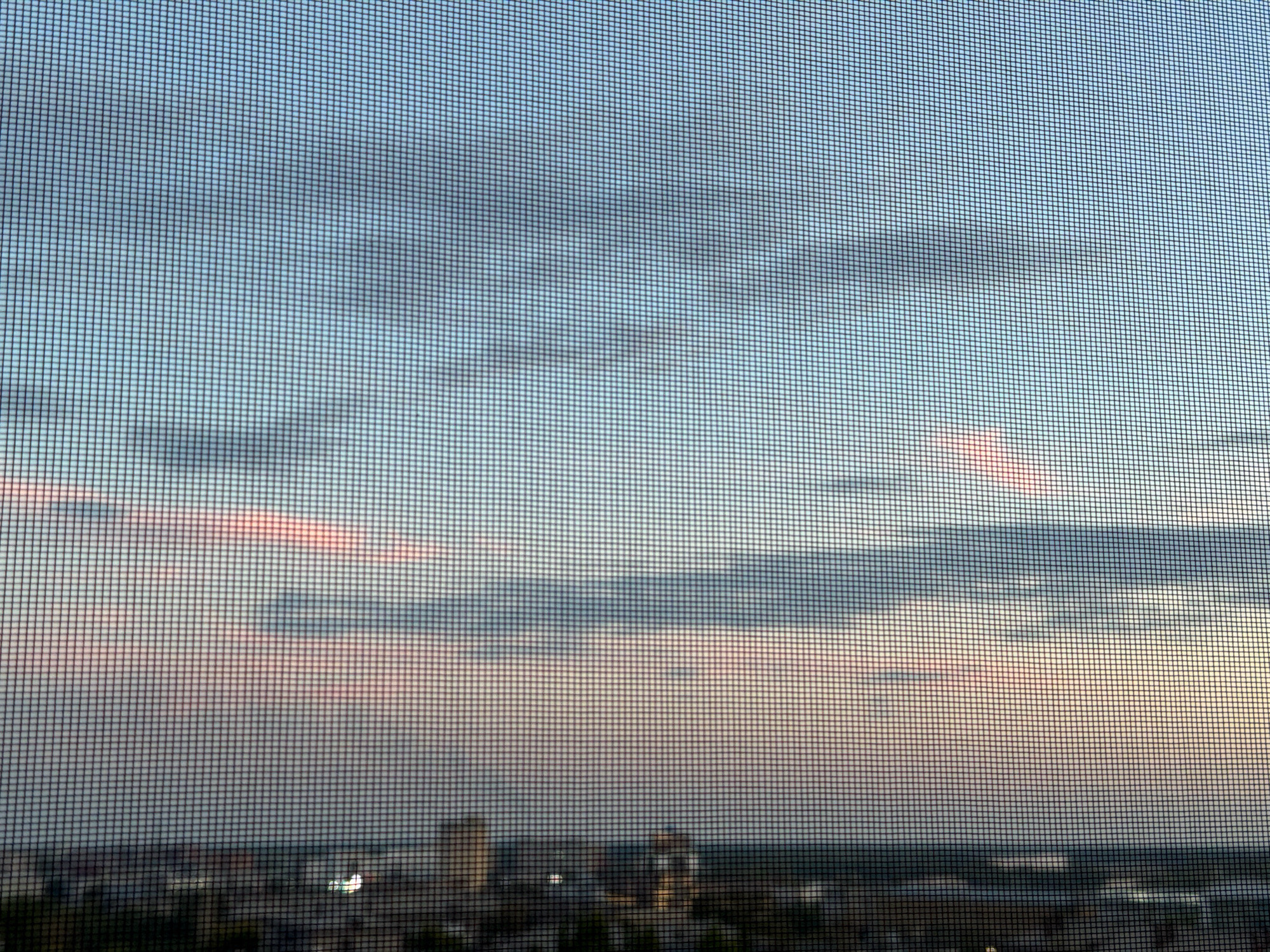The image depicts a sky with scattered clouds, tinged with pink and blue hues, seen through a window screen. Below the sky, a city skyline is visible, with buildings and lights suggesting an urban environment. The overall impression is that of a sunset or sunrise over a city. Without additional information or context, it is not possible to determine the specific location within Pavlodar, Kazakhstan.