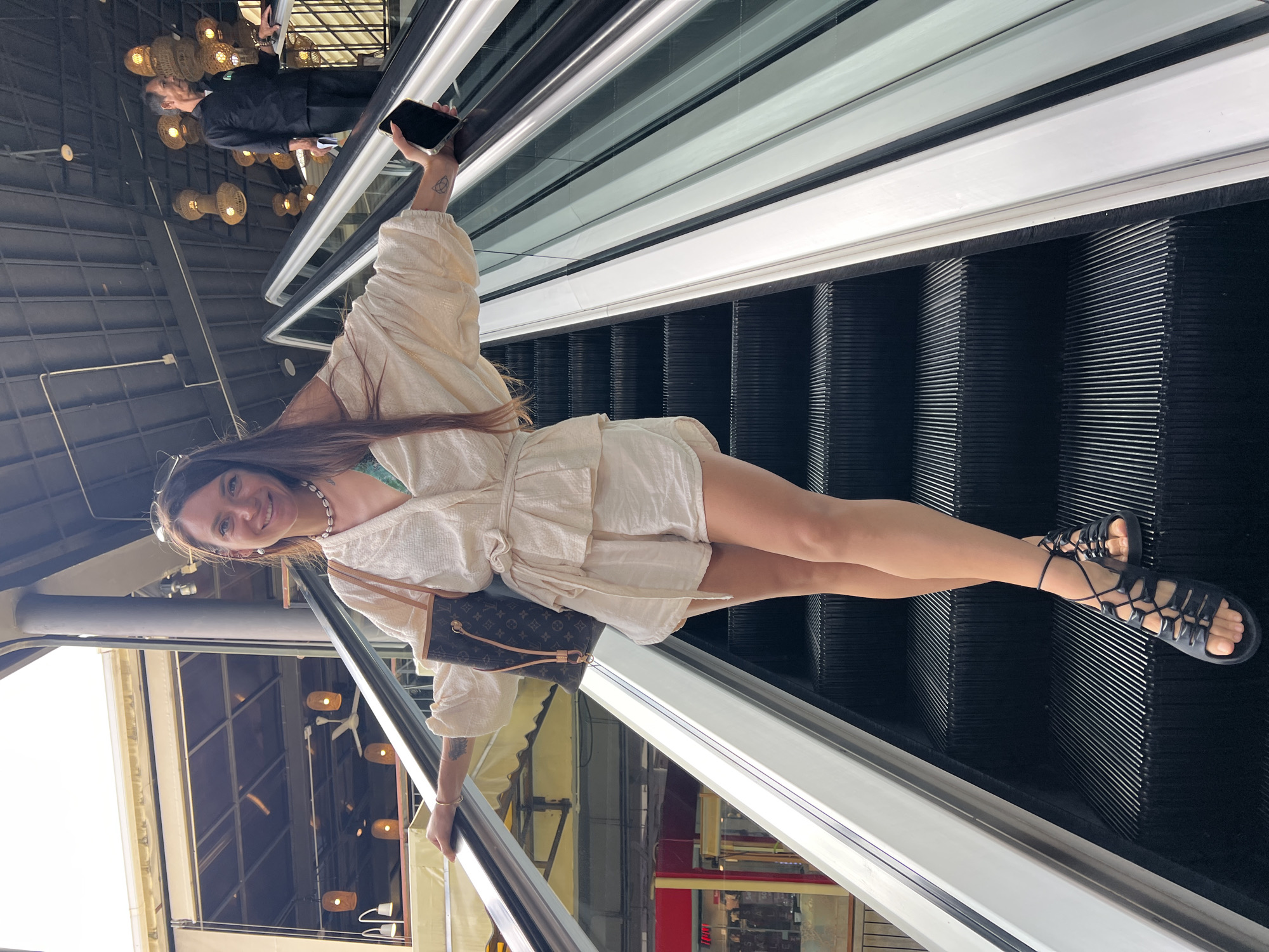 A woman is standing on an escalator, holding her phone in her right hand and her left arm resting on the escalator handrail. She is wearing a white, loose-fitting two-piece outfit and black sandals. She has a brown designer handbag slung over her left shoulder and is smiling at the camera. The escalator is moving upwards. In the background, there are lights hanging from the ceiling and another person in a black uniform is also on the escalator. The location is likely an indoor shopping center or a similar public building. It is not possible to determine the city of Ban Rak, Thailand from the image.