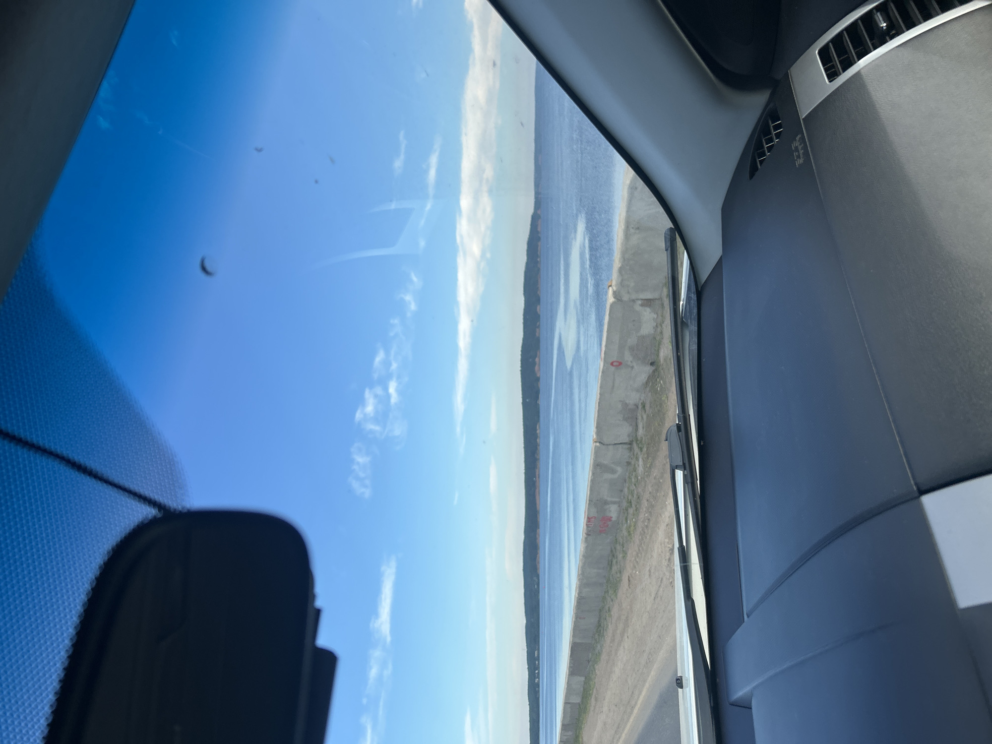 An interior view from a vehicle traveling on a road adjacent to a body of water. The foreground shows the dashboard and windshield of the vehicle, with a rearview mirror mounted at the top center. Through the windshield, a concrete barrier runs along the edge of the road, separating it from the calm, blue water. A distant, tree-covered shoreline forms the horizon under a bright blue sky with scattered white clouds. The scene appears to be on a sunny day, possibly near Chaikovsky, Russia, given the landscape and typical roadside infrastructure. There are no people or specific identifiable objects in the immediate vicinity.