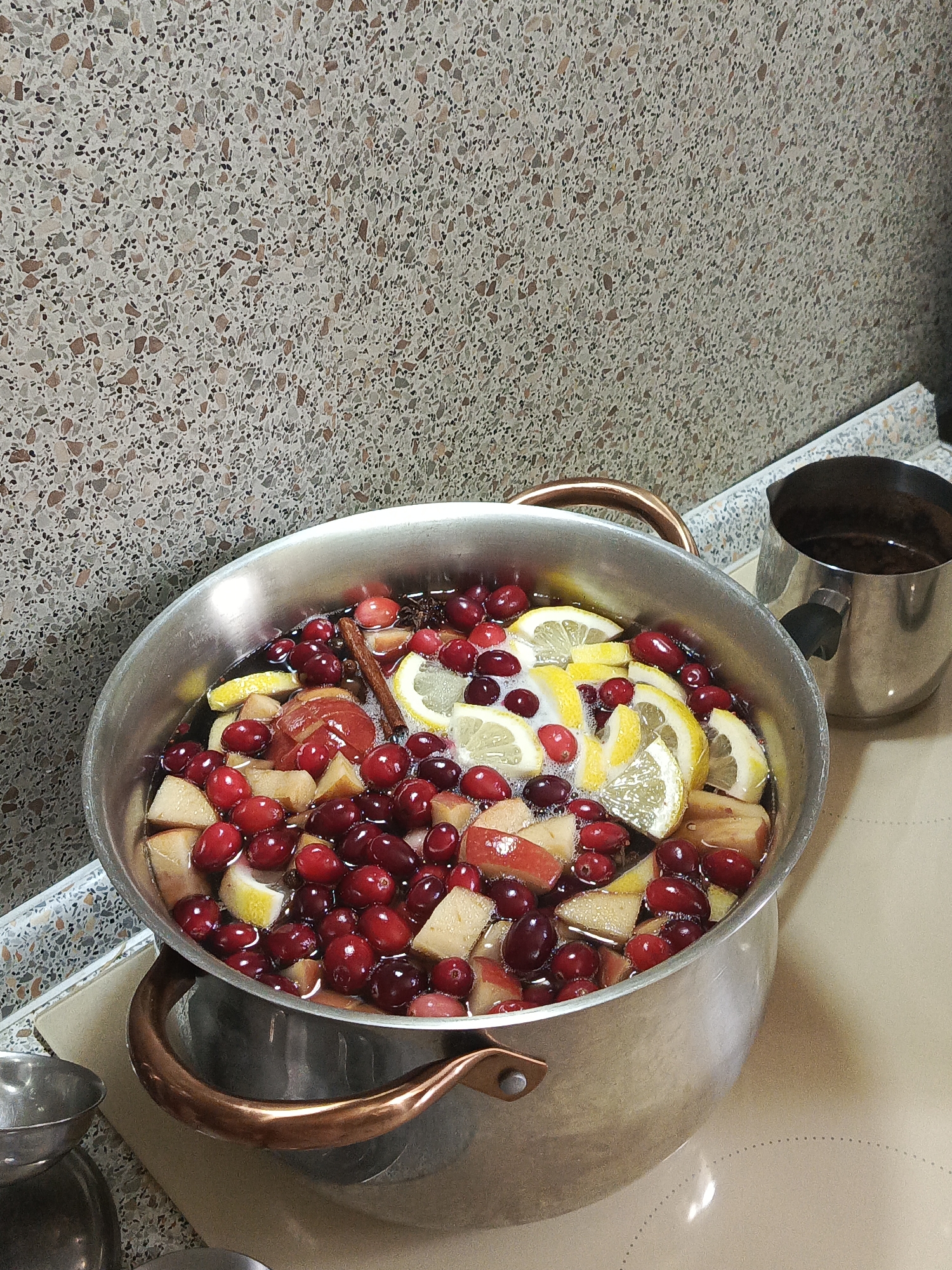 The image depicts a kitchen scene in the city of Северный, Russia. A large, cylindrical metal pot with two copper-colored handles occupies the central foreground, resting on a light beige stovetop. The pot is filled with a dark liquid containing an assortment of fresh fruits and spices. Visible ingredients include numerous whole, red cranberries, several yellow lemon slices, and irregularly cut pieces of apple or pear. A single brown cinnamon stick is submerged, and what appears to be a star anise pod is partially visible. The liquid surface shows a slight accumulation of foam.
To the right of the main pot, a smaller, dark metallic pitcher or pot with a black handle is positioned on the stovetop, containing a dark, undiscernible substance. The stovetop itself is a smooth, light beige surface with faint dotted circular patterns, indicating cooking zones.
Behind the pot, a wall is covered with a patterned material resembling terrazzo. This material consists of numerous small, irregular fragments of varying shades of grey, brown, and black embedded in a lighter, off-white or light grey matrix. A slight reflection is visible on the wall surface. In the bottom left corner, a portion of another small metal container is partially visible. The lighting suggests an indoor setting, likely artificial.