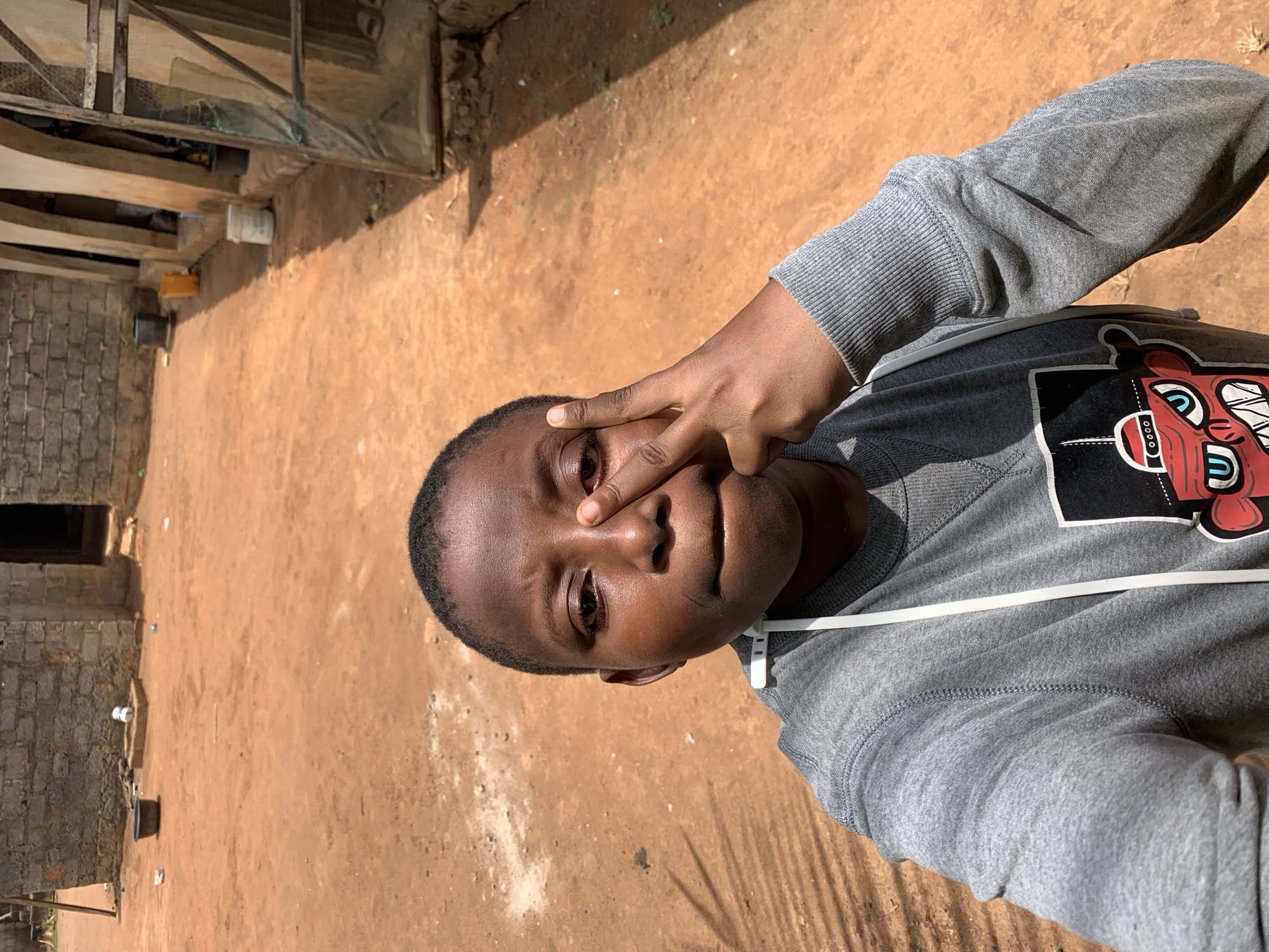 The image shows a person with dark skin, wearing a gray sweatshirt with a cartoon graphic, holding two fingers to their face. The background shows a reddish-brown dirt ground, and a building constructed of stone. Given the context of the prompt, the location is likely Ikorodu, Nigeria.