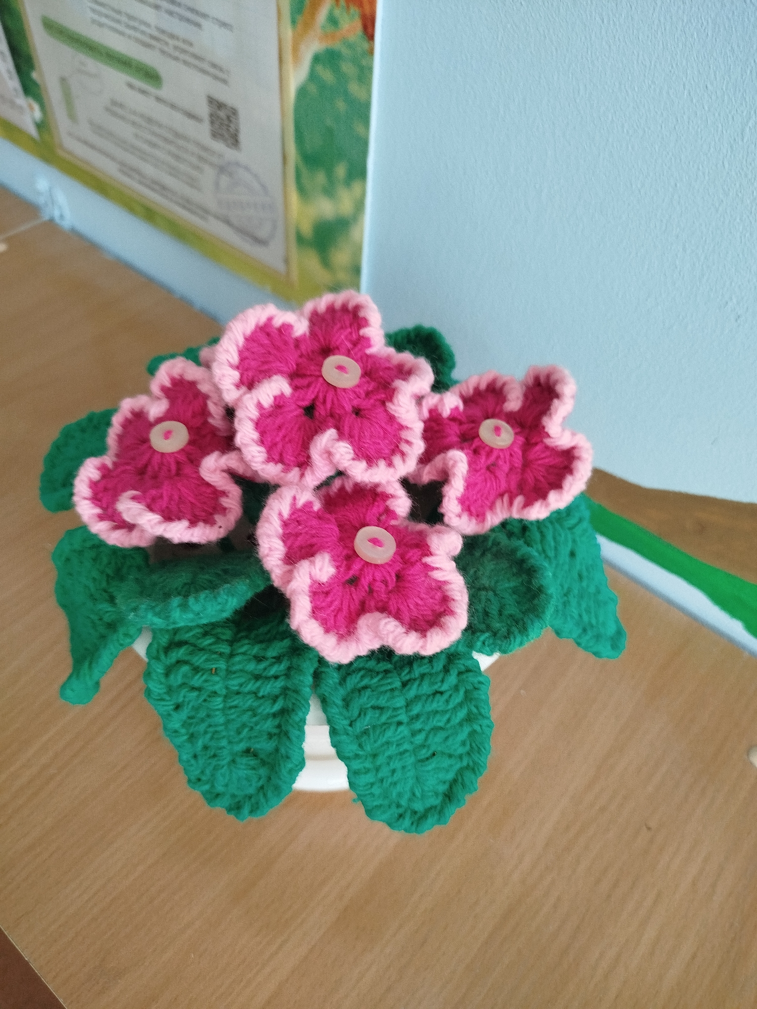 This media file shows a close-up of a crocheted potted plant with pink and light pink flowers and green leaves. The plant is placed on a wooden surface. In the background, there is a light blue wall and a bulletin board with some papers and a QR code. The lighting suggests it is daytime. The image appears to be taken indoors, likely in a room or classroom setting in Zavodoukovsk, Russia. There is no activity taking place; the focus is on the decorative crocheted item.
