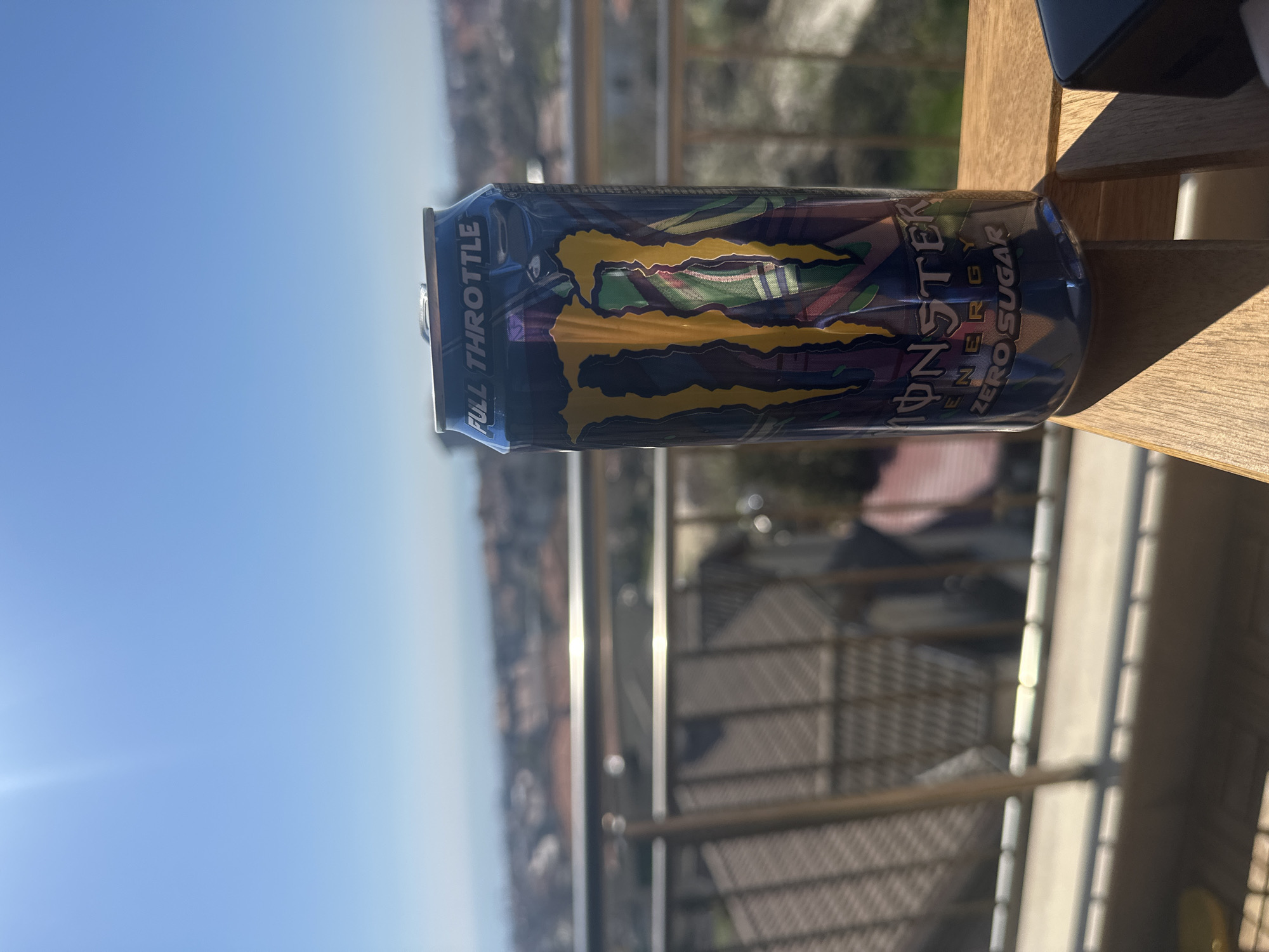 A tilted, close-up shot of a can of "Monster Energy Zero Sugar - Full Throttle" drink placed on a wooden surface. The background shows a cityscape with railings, suggesting a balcony or elevated position. The cityscape suggests it is an urban area with buildings and possible distant views of the sea or sky. Due to lack of clarity, exact location details cannot be precisely determined.