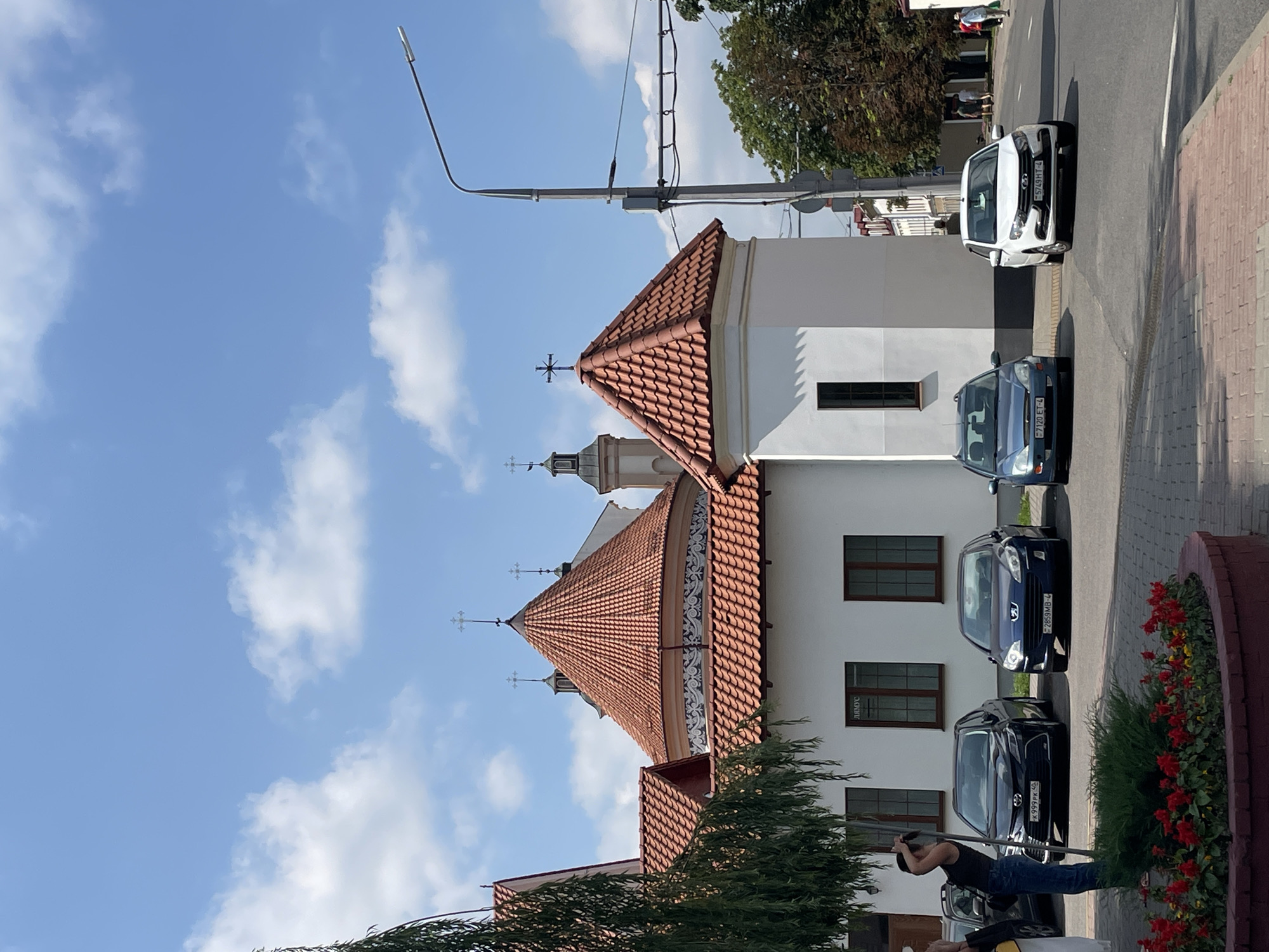 The image depicts a street scene in the city of Hrodna, Belarus, in front of the Holy Cross Church. Several cars are parked along the street. A woman in jeans and a dark top is standing near the curb, taking a photo with her phone. In the foreground, there is a circular planter filled with red flowers. The church features a white facade and red tiled roofs, with several crosses visible on its spires. A street light stands on the right side of the image. The sky is blue with scattered white clouds. In the background, other buildings and trees are visible, and a few people are walking on the sidewalk.