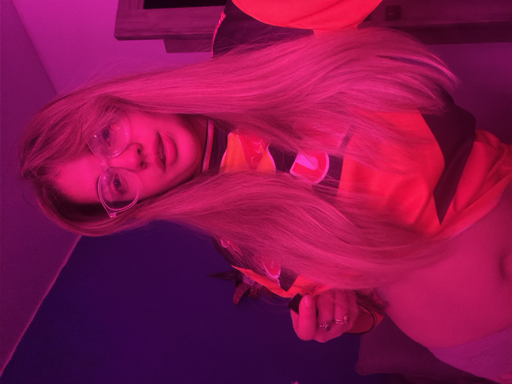 A young adult woman with long blonde hair and glasses is facing the camera. She's wearing a yellow and black jersey. The image is heavily saturated with pink and purple light. The setting appears to be indoors, but no location details are discernible beyond that. There is a dark object behind her. The woman's left hand is visible, holding an unidentifiable object. The lighting and composition suggest a casual, self-portrait style photograph. There is no evidence to suggest the photograph was taken in Большая Шильна, Russia.