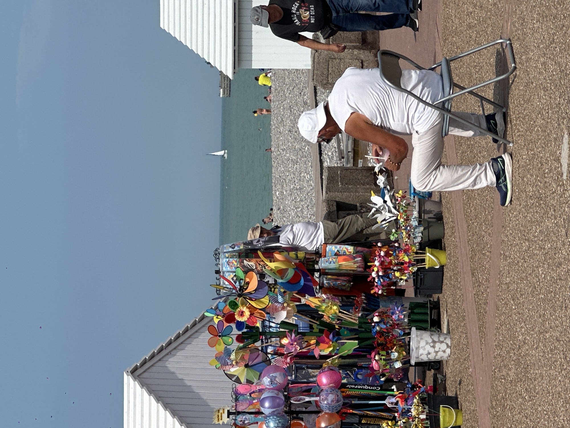 On a bright, sunny day in Dieppe, France, a man wearing a white t-shirt and white cap sits on a folding chair facing a stall of colorful pinwheels and toys. He holds a pink drink with a straw. The stall is laden with a variety of items, including small decorative birds, balloons, and an array of spinning pinwheels in various sizes and colors. Behind the stall, a man in a black t-shirt with "HARD ROCK IS NOT DEAD" printed on it walks past. The background features a pebble beach with a few people scattered across it, and the turquoise water of the English Channel. Two white beach huts are visible, one to the left and one to the right of the frame. A sailboat and a distant pier are visible on the water. The ground in the foreground is a mix of light-colored gravel and darker red brick paving.
