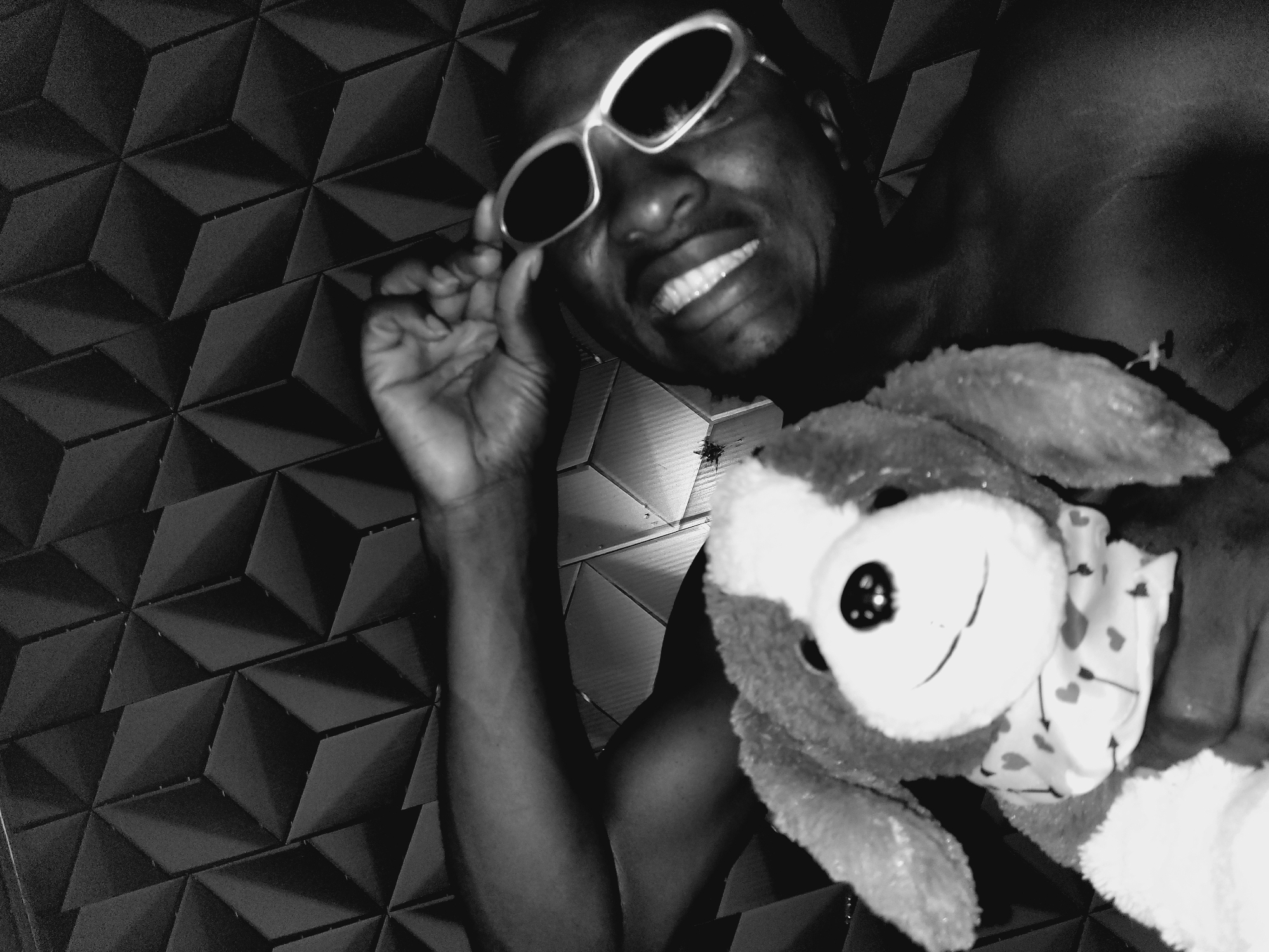 In Bukuru, Nigeria, a person with dark skin and a shaved head lies down, holding a stuffed dog. The person is wearing sunglasses and smiling broadly, with their hand adjusting the sunglasses. The background consists of a textured black wall. The stuffed dog is positioned in the foreground, obscuring a portion of the person's chest.
