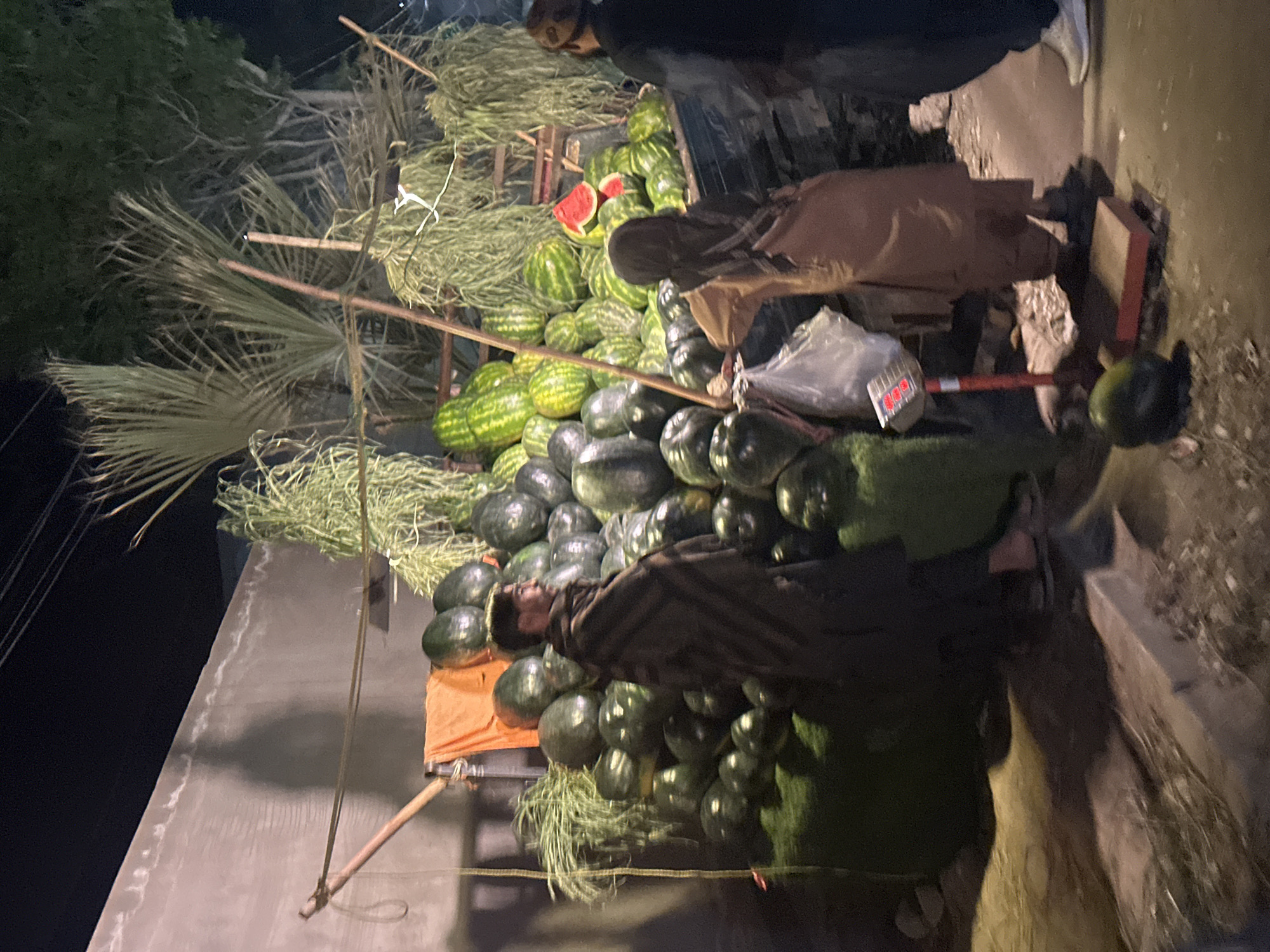 In Quetta City Tehsil, Pakistan, a street scene shows a watermelon stand at night. The stand is constructed of wooden supports, holding a large pile of green watermelons. Some watermelons are dark green and some are striped green. A couple of watermelon slices are visible. The stand is decorated with dried grass. Two people are interacting with the stand, one wearing a brown coat and one wearing a black jacket and mask. A scale is on the ground near the stand. A curb is on the right. A wall is on the left. A palm tree is nearby.