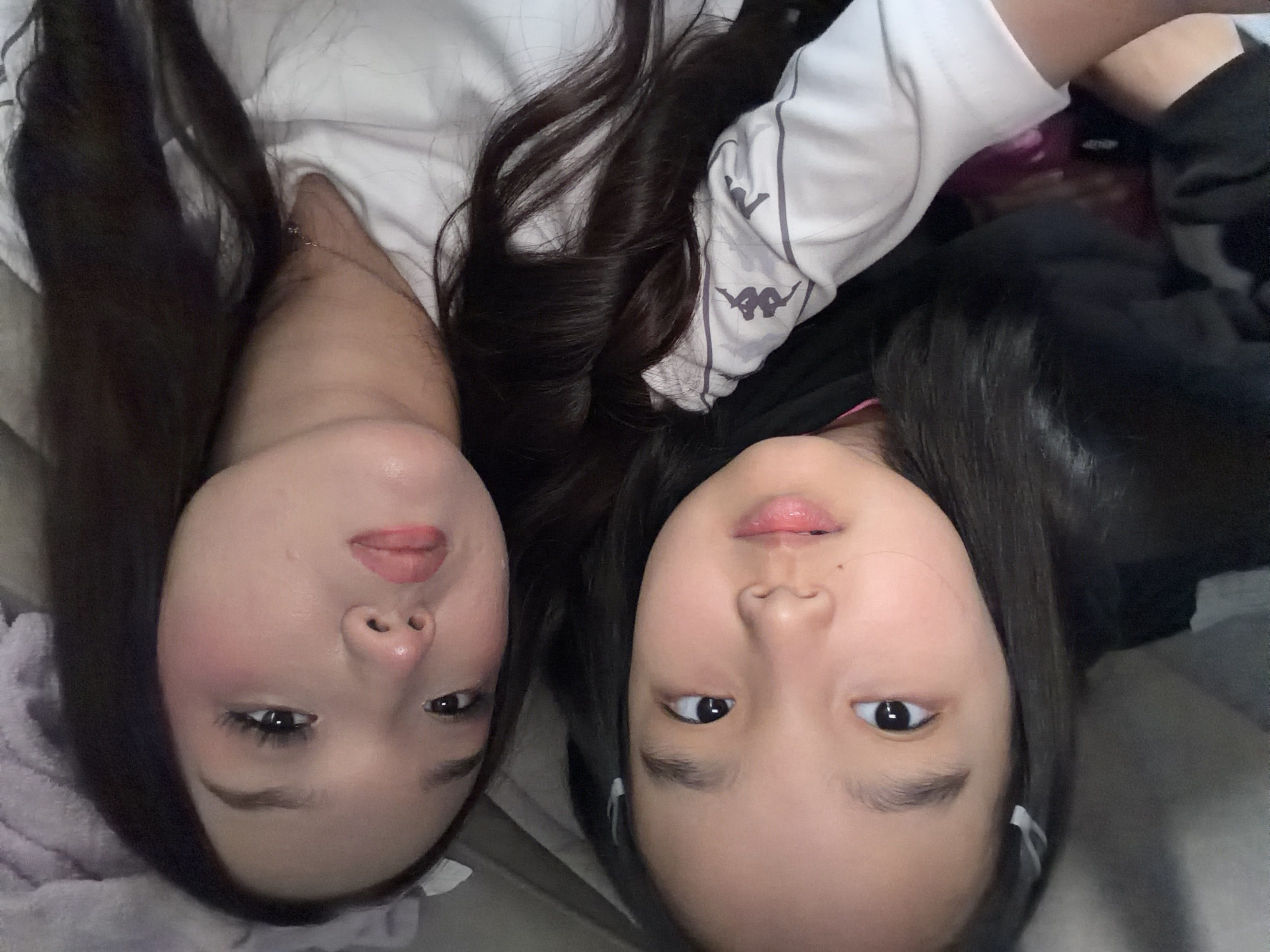 Two young females are captured in a close-up selfie-style photograph, their faces dominating the frame. The female on the left has dark hair and appears to be younger, with a light complexion and small hair clips visible. The female on the right has dark, wavy hair and is wearing makeup, including noticeable eyelashes and blush. She is looking directly at the camera, while the younger female's gaze is slightly averted. Both are wearing casual clothing. The background is blurred, suggesting an indoor setting, possibly a home environment. There is no indication of the location within a city or country, and therefore no information about "Unknown, Russia" can be provided. The image focuses solely on the two individuals and their upper bodies.