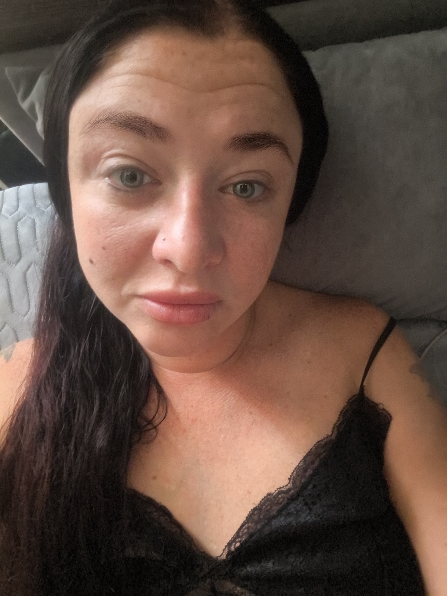 A close-up, indoor, selfie-style photograph shows a woman's face and upper body. She has dark hair that is pulled back, revealing her forehead and eyebrows. Her eyes are open, and she is looking towards the camera. Her nose is slightly turned, and her lips are slightly parted. She has a mole on her left cheek and a small stud piercing on her nose. She is wearing a black lace camisole. The background appears to be a couch or upholstered furniture. The lighting is natural and appears to be coming from the right side of the frame. The image was taken in Lisakovsk, Kazakhstan.