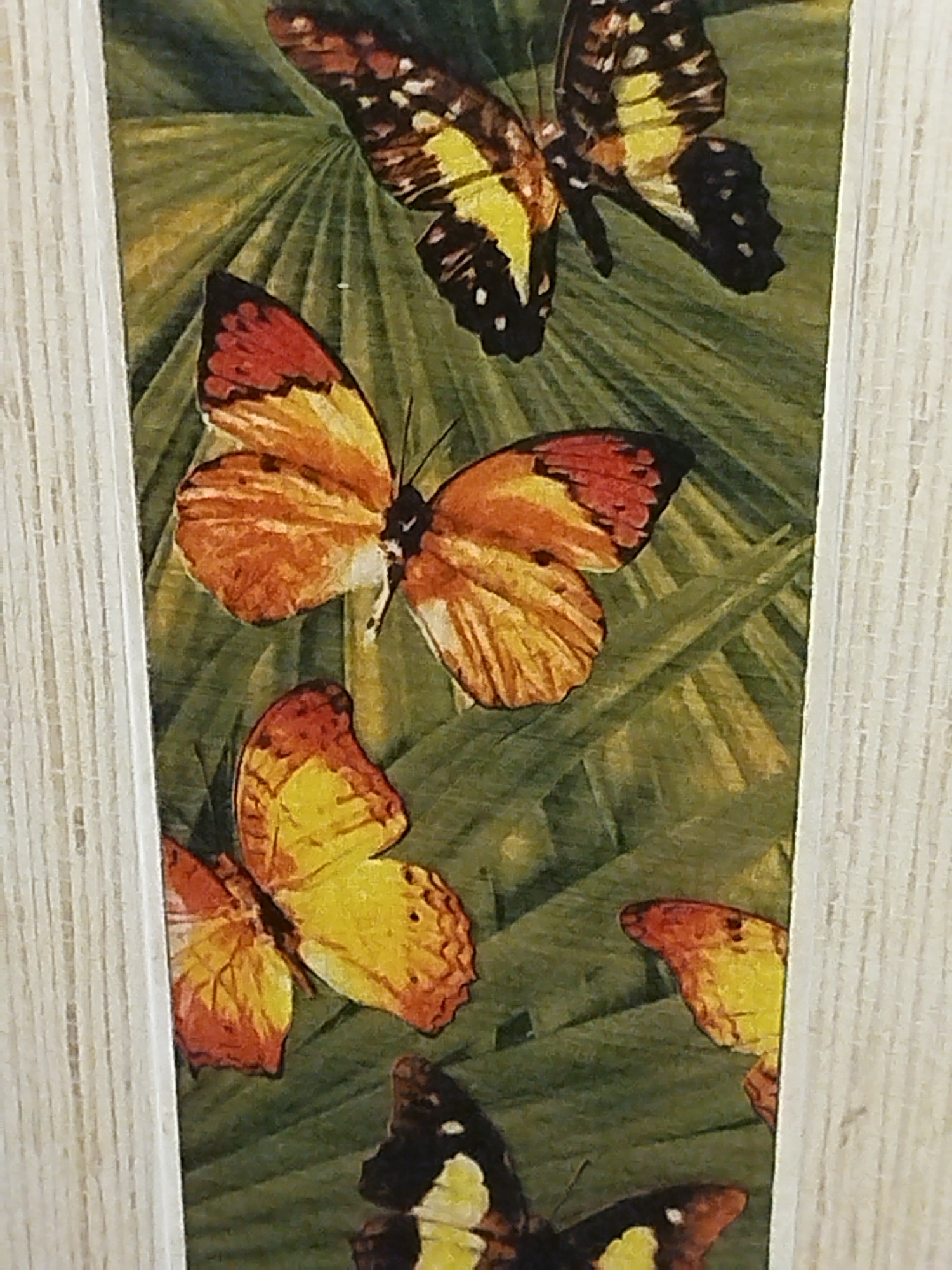 This is a close-up, vertical, eye-level, interior shot of a framed decorative panel featuring a painted depiction of several butterflies against a green, leafy background. The panel is framed by a light-colored, textured material, possibly wood. From top to bottom, the butterflies are: one with black, yellow, and white markings; one with orange and yellow wings; another with yellow and orange wings; and a partial view of a butterfly with black, yellow, and white markings at the bottom. The painting style is realistic. The location of Leninsk-Kuznetsky, Russia, is not discernible from the image.