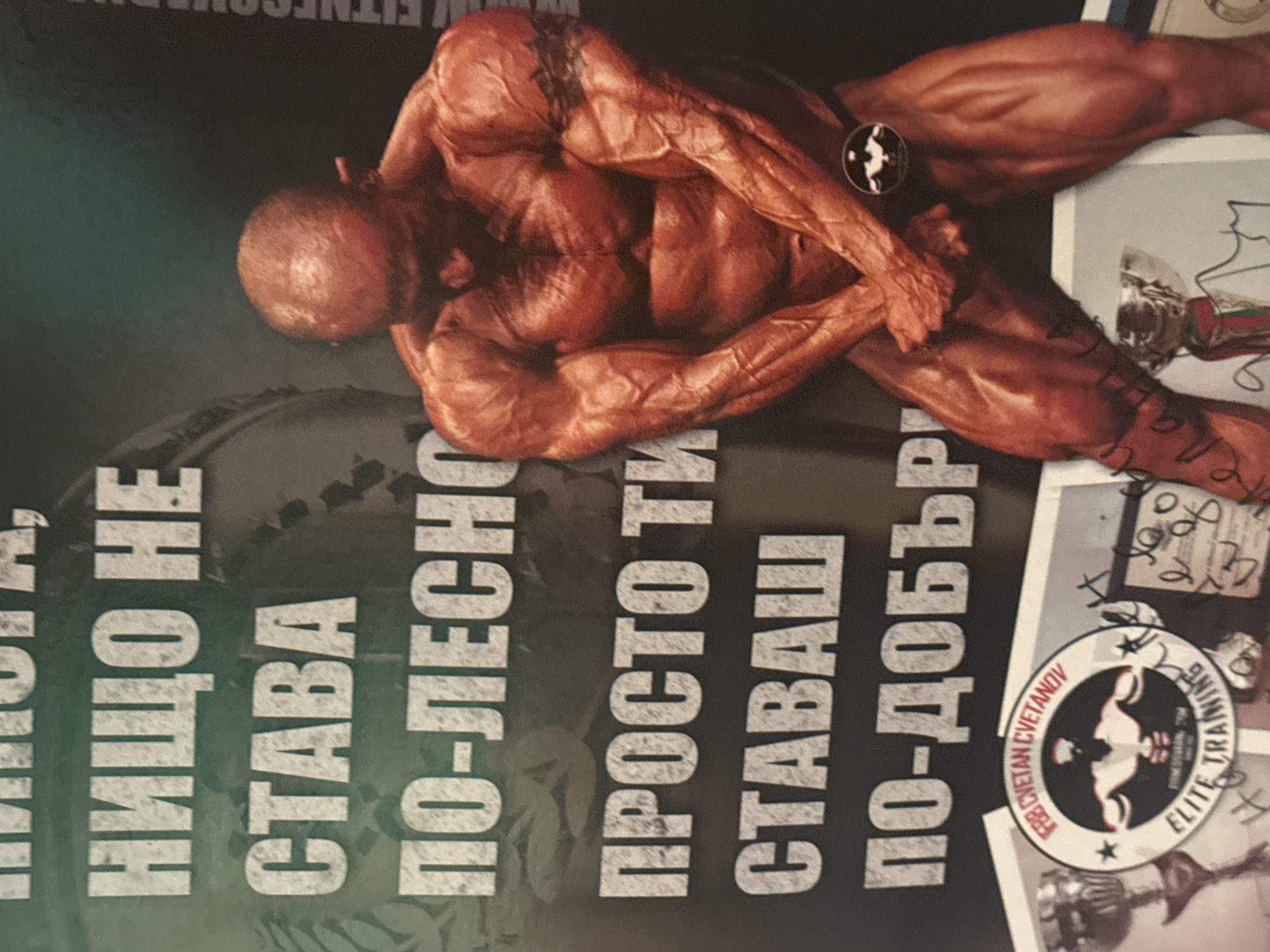 The image displays a highly muscular, bald bodybuilder, heavily spray-tanned, striking a pose that emphasizes his back and arm musculature. He is positioned against a dark, faded green and black background, which appears to be a promotional poster, magazine page, or advertisement. His physique is exceptionally defined, showcasing prominent veins and muscle separation, indicative of intense training and competition.
To the left of the bodybuilder, large white Cyrillic text is arranged vertically. The text reads: "НИЩО НЕ СТАВА ПО-ЛЕСНО ПРОСТО ТИ СТАВАШ ПО-ДОБЪР," which translates to "NOTHING BECOMES EASIER YOU JUST BECOME BETTER." A circular logo is visible in the lower right, featuring a stylized depiction of a bodybuilder's head and shoulders. Around the logo's perimeter, text reads "FB2 CVETAN CVETANOV" and "ELITE TRAINING," which is repeated. Partially visible at the top is a website address beginning with "WWW.FITNESCOM". In the background, behind the bodybuilder's legs, faint outlines of what appear to be trophies or awards are visible, along with some indistinct handwritten marks or autographs. The overall scene suggests a focus on professional bodybuilding and elite fitness training, likely originating from Varna, Bulgaria, given the Cyrillic text and provided context.