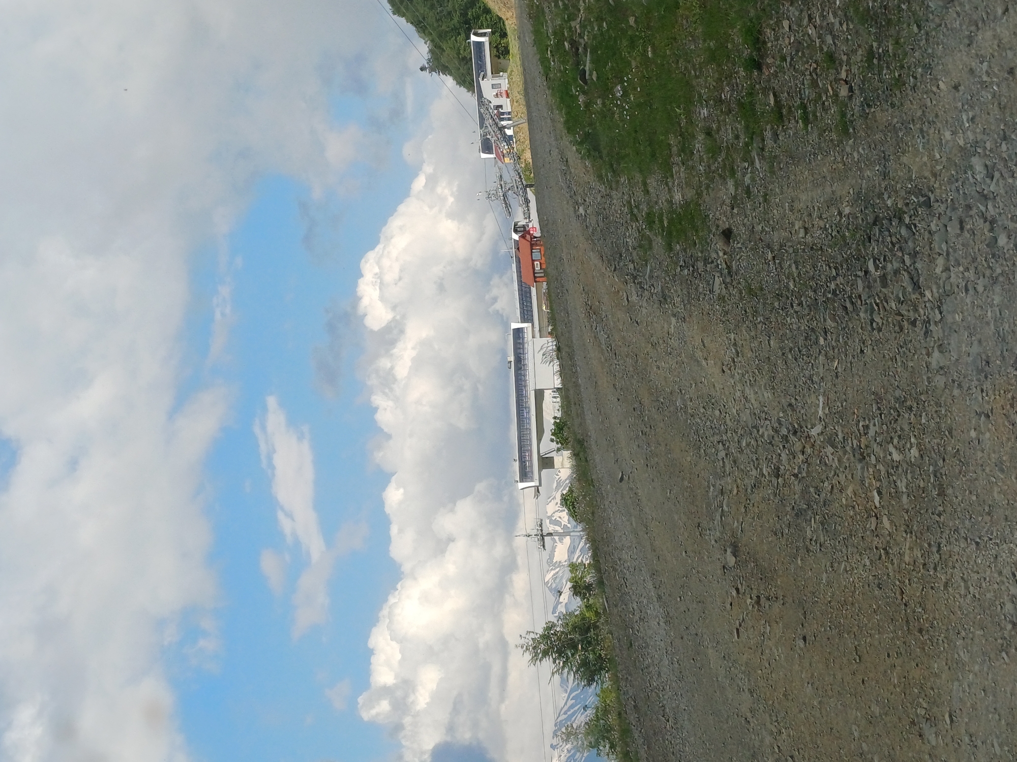 The image depicts a mountainous landscape with a ski lift and buildings visible on a slope. A gravel road leads upwards in the foreground. The sky is partly cloudy with blue patches and large white cumulus clouds. In the background, snow-capped mountains can be seen behind the ski lift infrastructure. The ski lift appears to be a modern system with enclosed cabins. The overall scene suggests a resort area in a mountainous region.