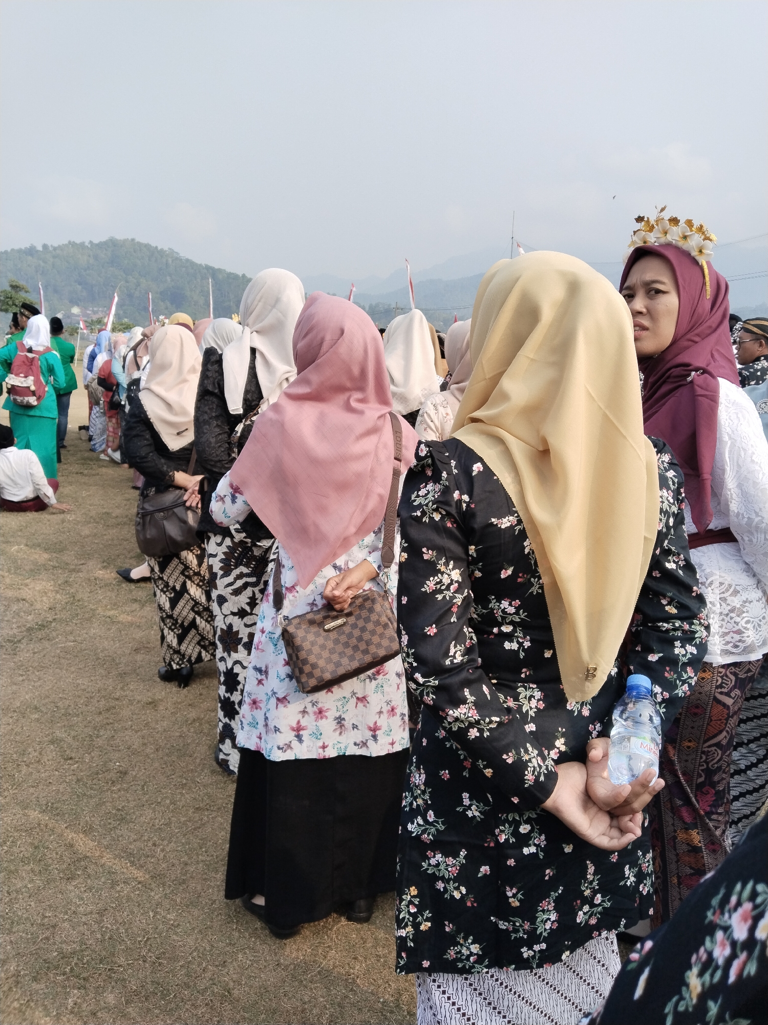 The image depicts a line of people, primarily women wearing hijabs and traditional Indonesian attire, in Munjungan, Indonesia. The foreground focuses on the backs of several women. One woman in a floral print black blouse and a light yellow hijab is holding a bottle of water. To her left, another woman in a floral print white blouse and a pink hijab has a checkered brown purse. Further to the left, a woman in a patterned black and white batik skirt and a white hijab is visible. In the background, more individuals are seen in the distance, and a hilly landscape with some buildings and trees can be observed. The ground is covered in dry grass. The sky is overcast.