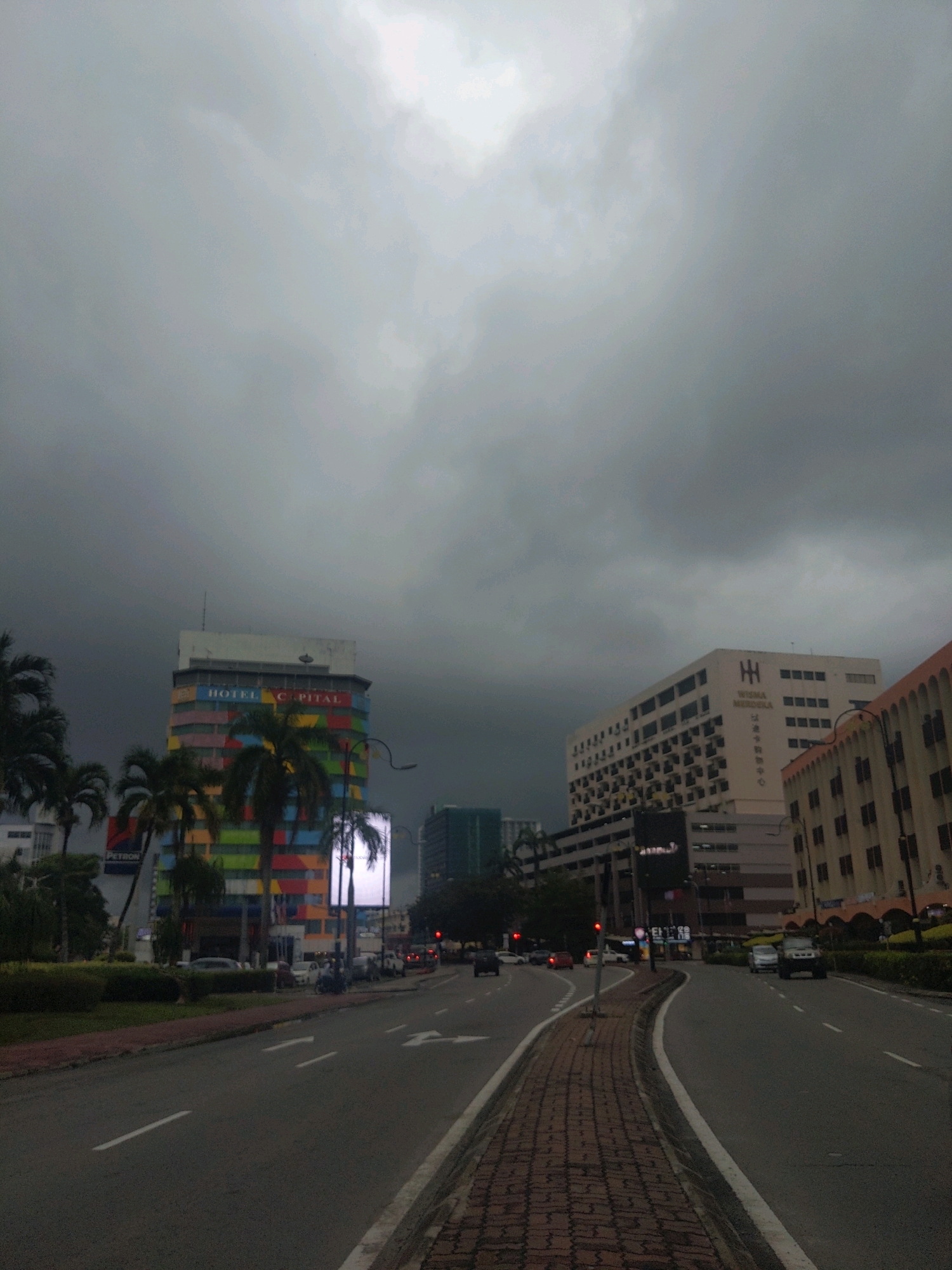The image shows a street scene in Kota Kinabalu, Malaysia under a dark, cloudy sky. A multi-colored building, identified by signage as "HOTEL CAPITAL," stands prominently on the left. Adjacent to it is a building with the text "WISMA MERDEKA." The street is paved with asphalt, and there is a brick sidewalk. Several cars and a few motorcycles are visible on the road, indicating moderate traffic. Lush green vegetation and palm trees line the sides of the road. The overall atmosphere is somber, possibly indicating impending rain or a recent downpour.