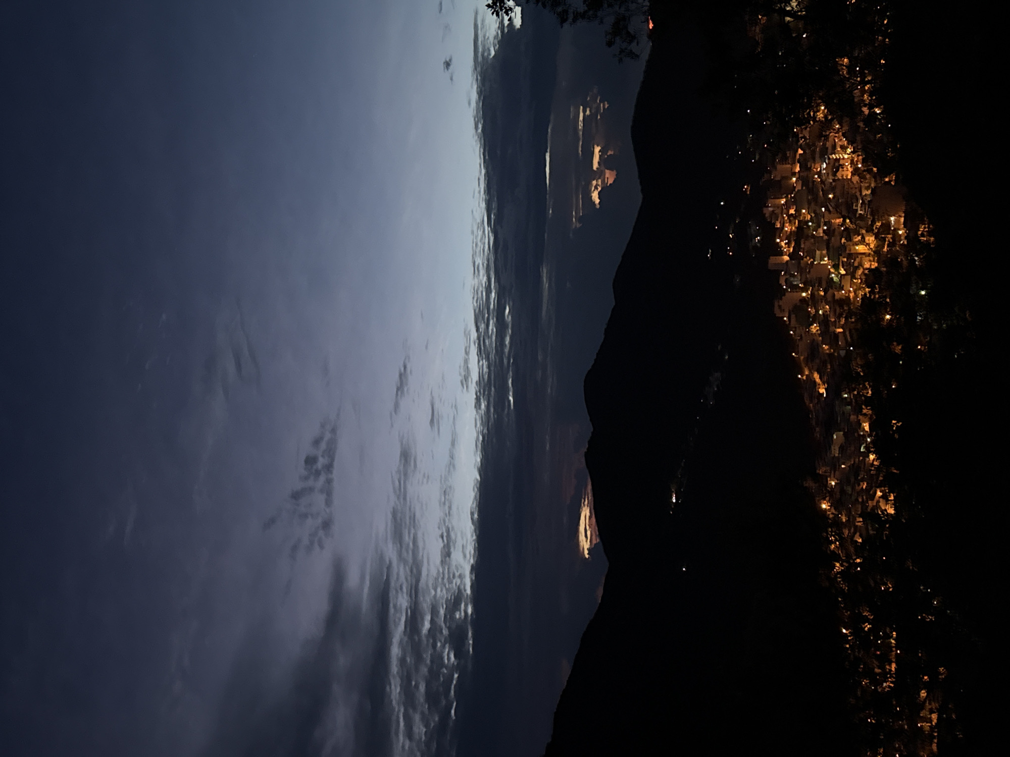 At dusk in Bac Nha Trang Ward, Vietnam, a dark mountain range forms a silhouette against a sky transitioning from deep blue to lighter shades with streaks of clouds. The city below is illuminated by a scattering of yellow lights, suggesting a dense urban area extending into the darkness.