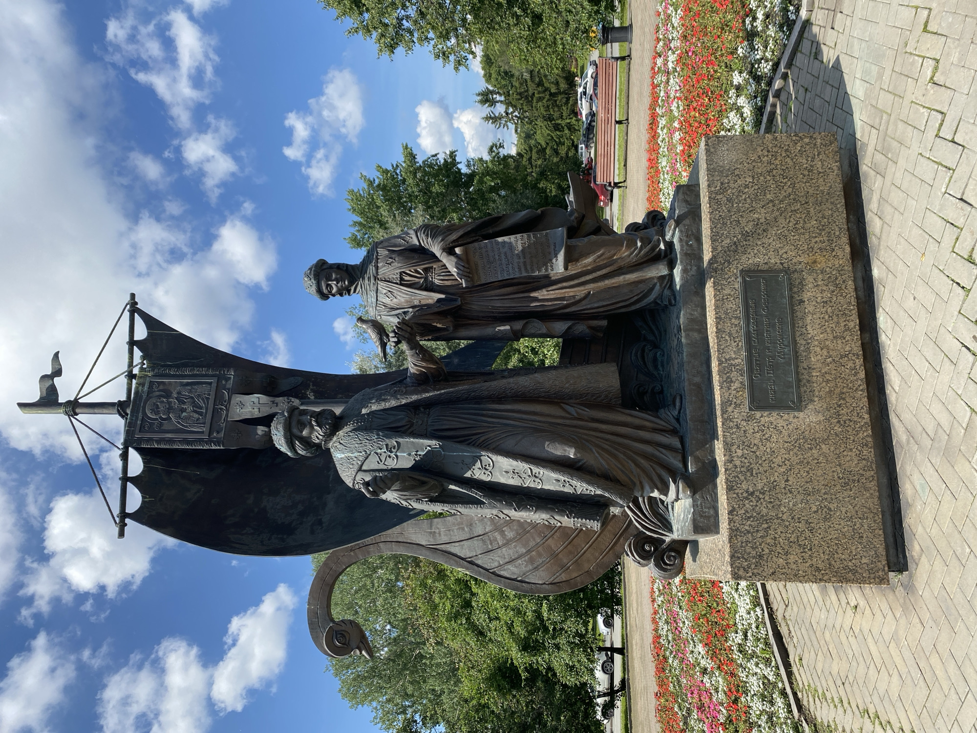 The photograph shows a bronze statue in a park setting. The statue depicts two figures, likely historical figures, standing next to a large, stylized sail and ship prow. One figure is holding a bird, while the other holds a scroll. The base of the statue is made of granite and has a plaque with an inscription. In the background, there are trees, a flowerbed with colorful flowers, a paved walkway, and a park bench. The sky is blue with scattered clouds.
The statue is located in Yekaterinburg, Russia, and depicts Saints Cyril and Methodius, the creators of the Glagolitic alphabet.
