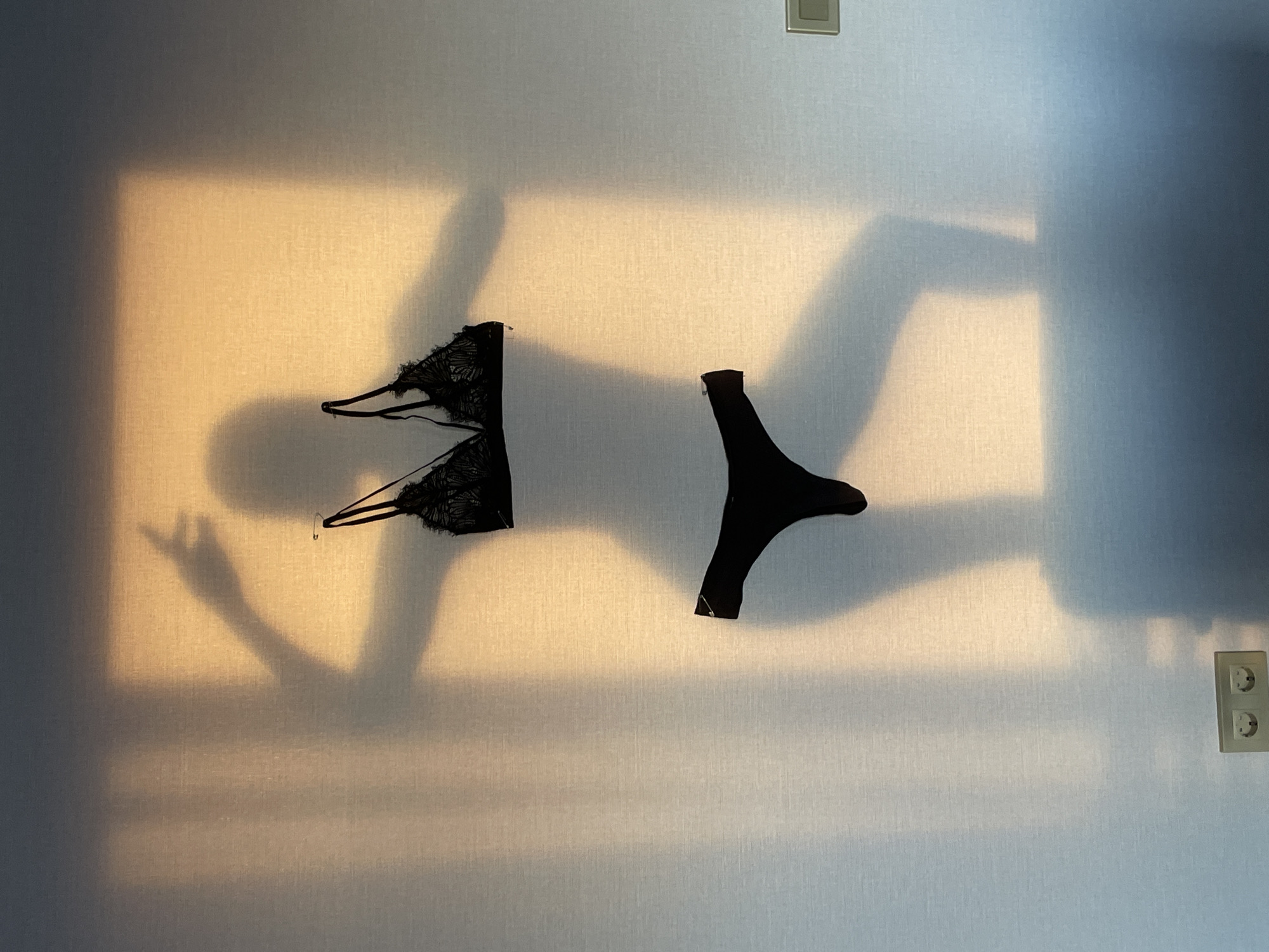 The image shows a brightly lit wall in what appears to be an interior setting. Affixed to the wall are two black lingerie items: a bra on the left and a thong on the right. The wall also features the shadows of a person's head, body, and arms, and a rectangular light fixture. Additionally, there are electrical outlets visible on the lower right of the wall. The background is textured and has two separate colors due to the brightness. The image contains a light switch. Based on the instructions that it should be located in the city of Подшивалово, Russia, this image contains the information that it is located there.