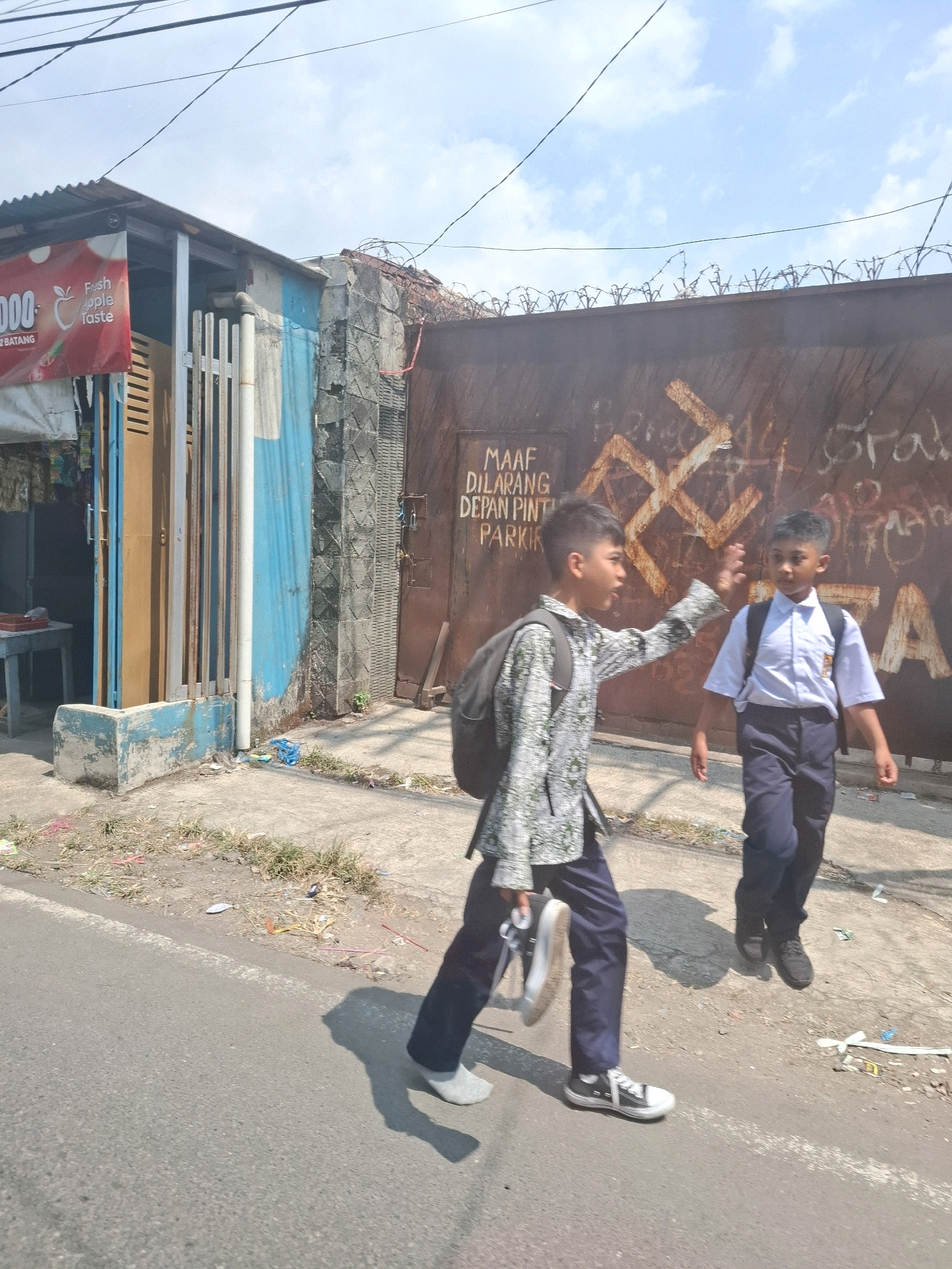The image depicts two young boys walking on a street in Sukabumi, Indonesia. The boy on the left, wearing a patterned shirt and dark pants, is carrying a backpack and a pair of black sneakers in his hand. He is extending his right hand towards the other boy. The boy on the right, wearing a white collared shirt and dark pants, is also carrying a backpack and walking ahead. Both boys appear to be students. In the background, there is a rusty metal gate with graffiti on it, including what appears to be a swastika symbol and text in Indonesian, "MAAF DILARANG DEPAN PINTU PARKIR" (Sorry, prohibited in front of the parking door). To the left of the gate is a small structure with a corrugated metal roof and blue and white painted walls, possibly a shop or a building entrance. There are barbed wires on top of the gate. The ground is paved and there is some debris scattered around. The sky is clear with some clouds.