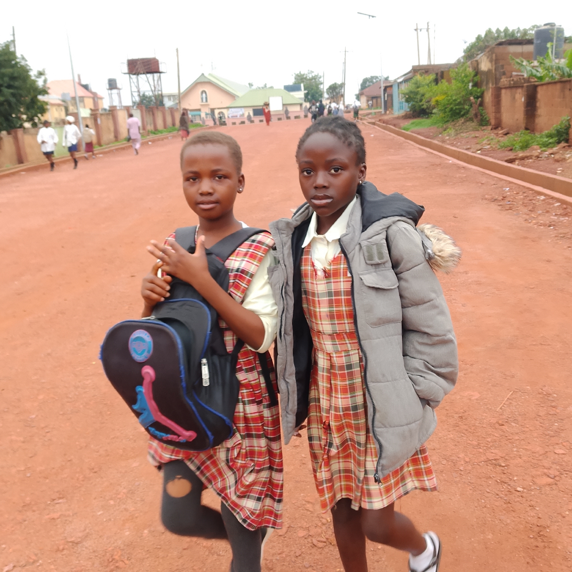 Two young girls in school uniforms are standing on a dirt road in Gura Top, Nigeria. The girl on the left is wearing a red and white plaid dress and a white top, carrying a black backpack. The girl on the right is wearing a similar plaid dress and a white top, with a grey puffer jacket over it. They appear to be posing for a photograph. In the background, other people and buildings are visible, suggesting a residential or semi-urban area. The road is made of reddish-brown dirt, and there is vegetation and some infrastructure like water tanks and utility poles in the distance. The sky is overcast.