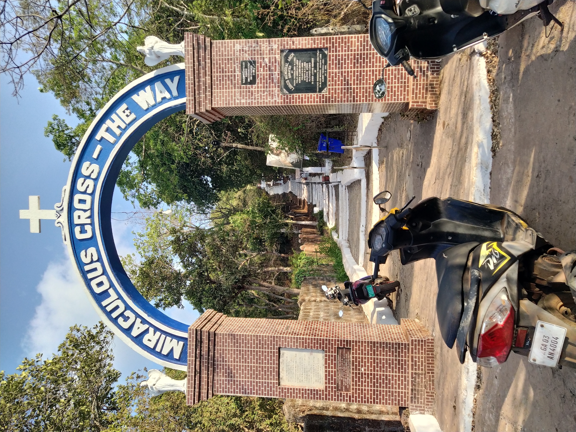 The image depicts the entrance to a cemetery in Assagao, India, marked by an arched blue sign that reads "MIRACULOUS CROSS - THE WAY". The sign is supported by two brick pillars, each topped with a white statue. Each pillar has a plaque. A dirt road leads from the entrance into the cemetery, with rows of white and gray tombs visible in the distance. Two motor scooters are parked near the entrance, partially obscuring the view. Trees surround the cemetery. The sky is blue, with some clouds visible on the left side of the image.